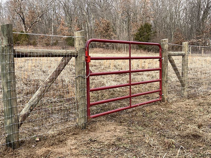 Red metal gate in a wooden fence at the edge of a dry field with bare trees behind it.
