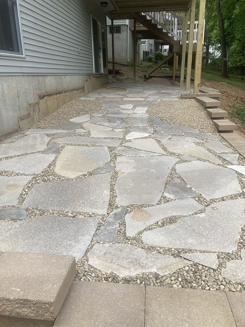 Stone walkway beside a house, with uneven paving and gravel under a covered porch.
