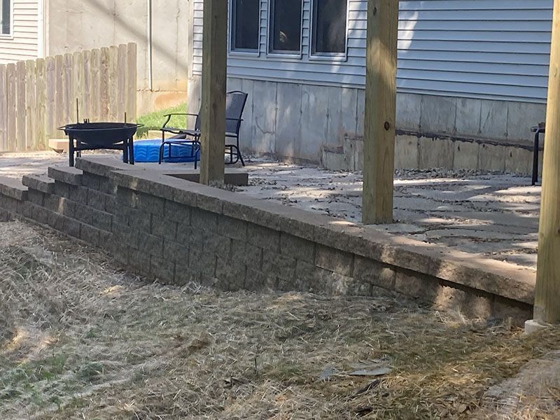 Backyard patio with stone retaining wall, wooden posts, and a small grill near a house.