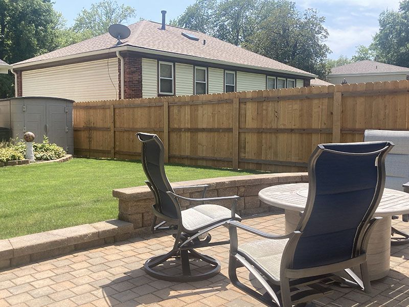 Backyard patio with two chairs and a small table on a brick paver area, beside a fenced lawn.