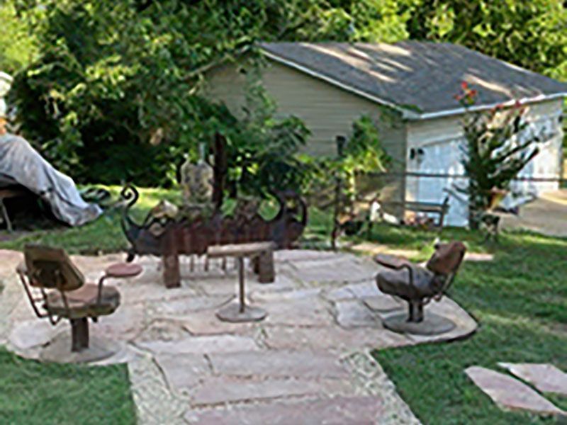 Backyard stone patio with metal chairs and fire pit beside a garage and trees