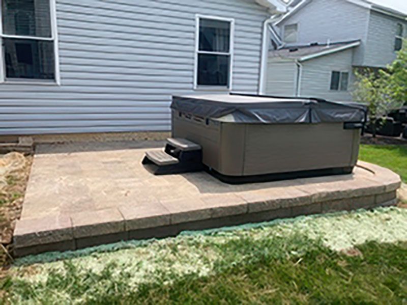Covered hot tub on a stone patio beside a house with grass in the foreground.