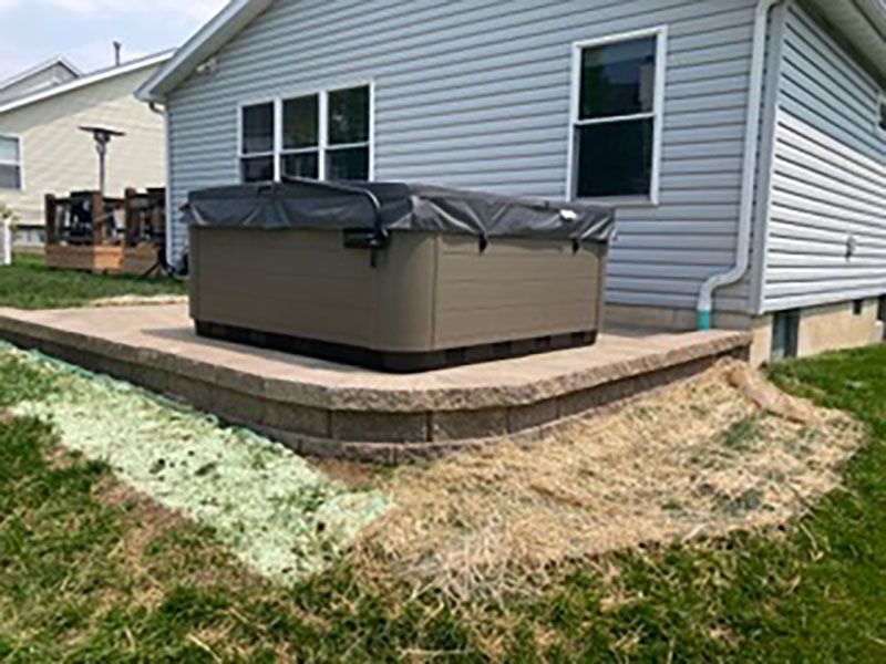 Covered hot tub on a raised patio beside a gray house, with a grassy yard and stone retaining wall.