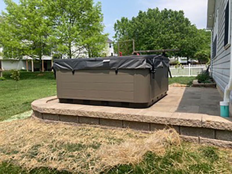 Covered hot tub on a raised patio in a backyard with trees and a house.