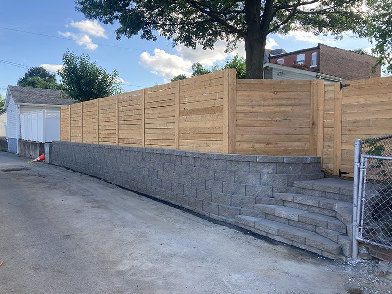 Newly built wooden fence atop a gray retaining wall along a residential driveway under a tree.