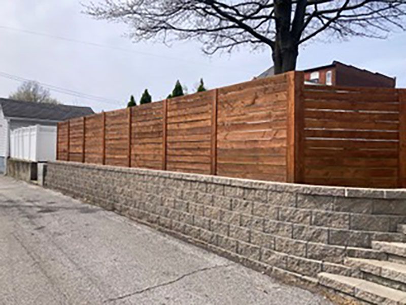Wooden privacy fence atop a stone retaining wall along a residential street with stairs at the right.