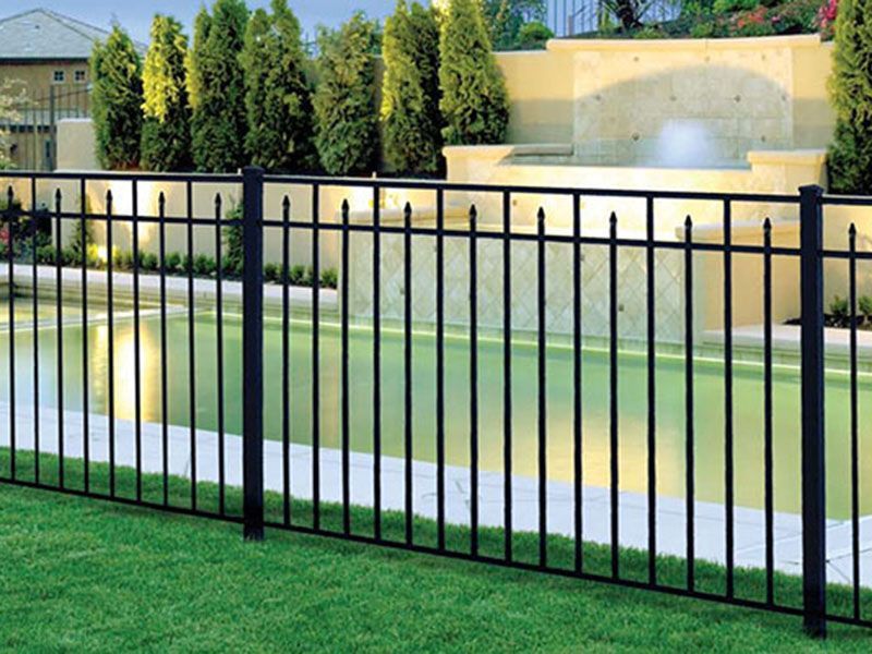 Black metal fence in front of a backyard pool and patio with green lawn and beige walls