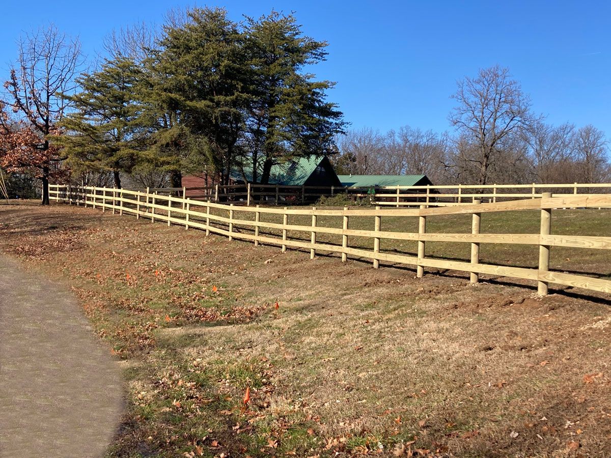 Wooden fence along a leaf-strewn path beside grassy fields and trees under a clear blue sky