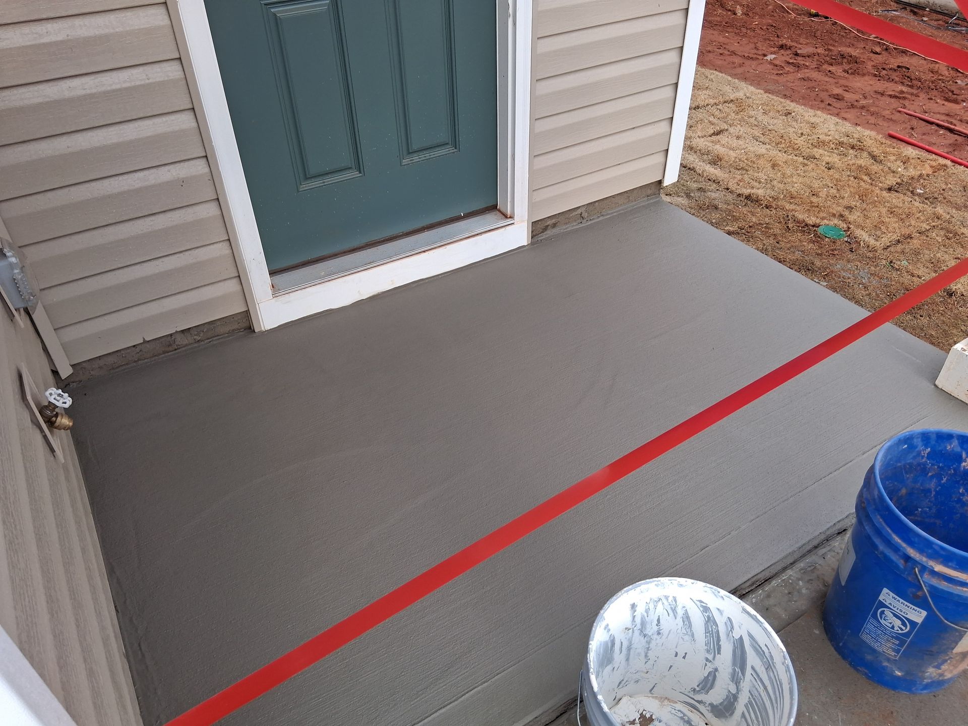 Freshly poured gray concrete porch step outside a house with blue siding and a green door, with buckets nearby.