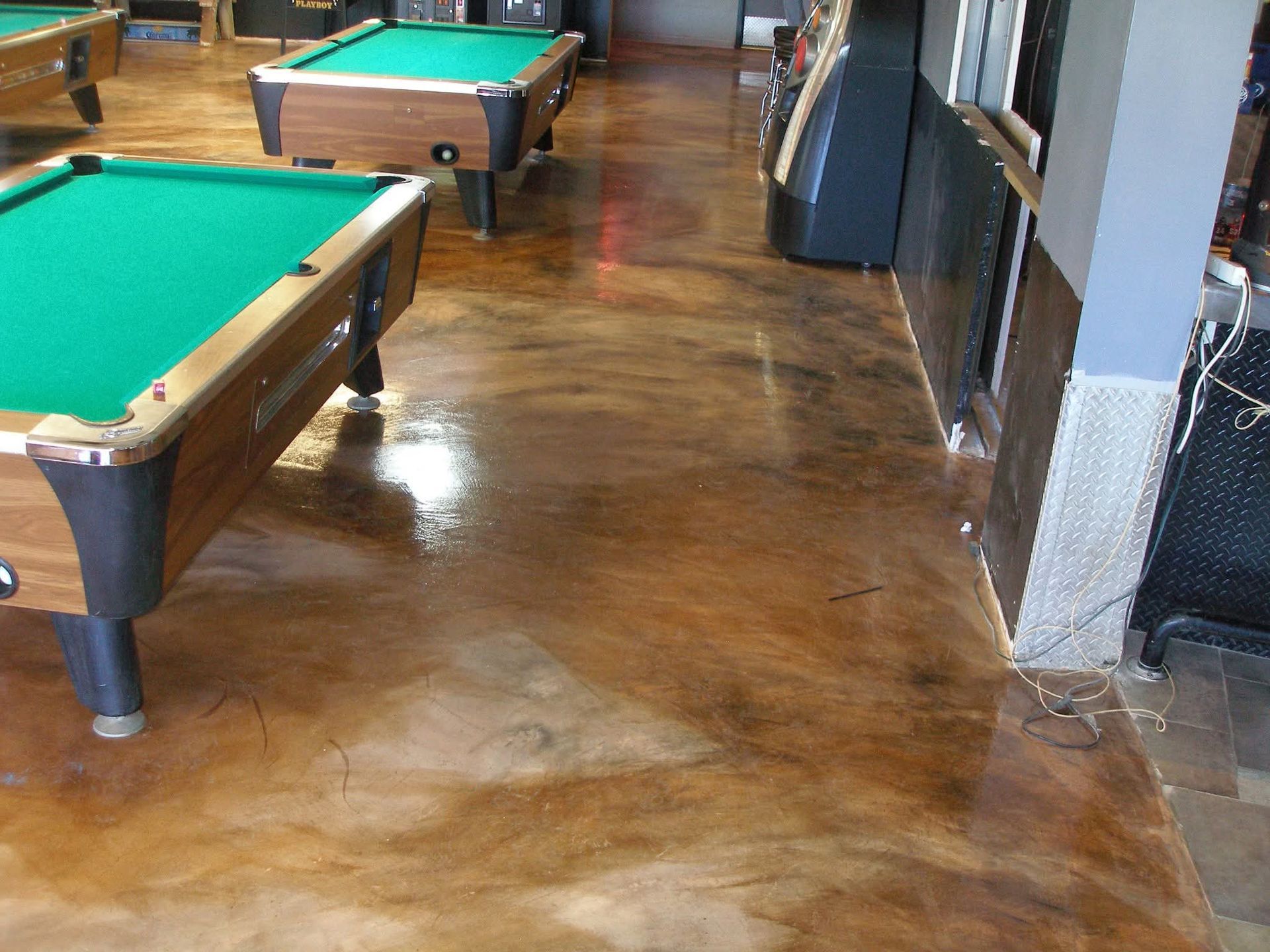 Billiard tables on a shiny, brown stained concrete floor inside a room.