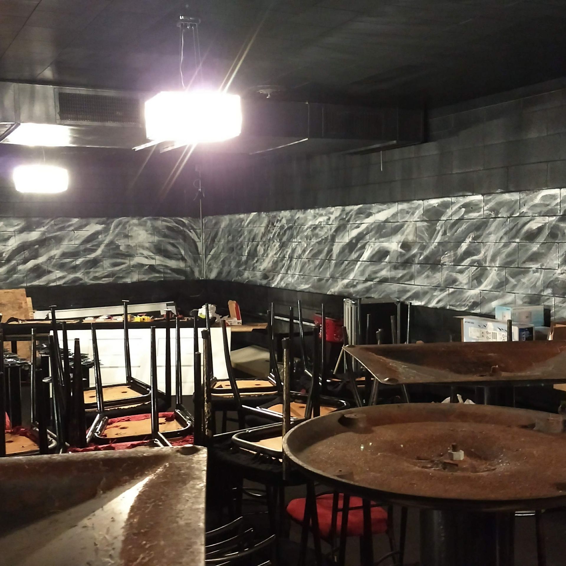A dimly lit room with stacked chairs, tables, and a wall featuring a black-and-white marbled, cloudy texture.
