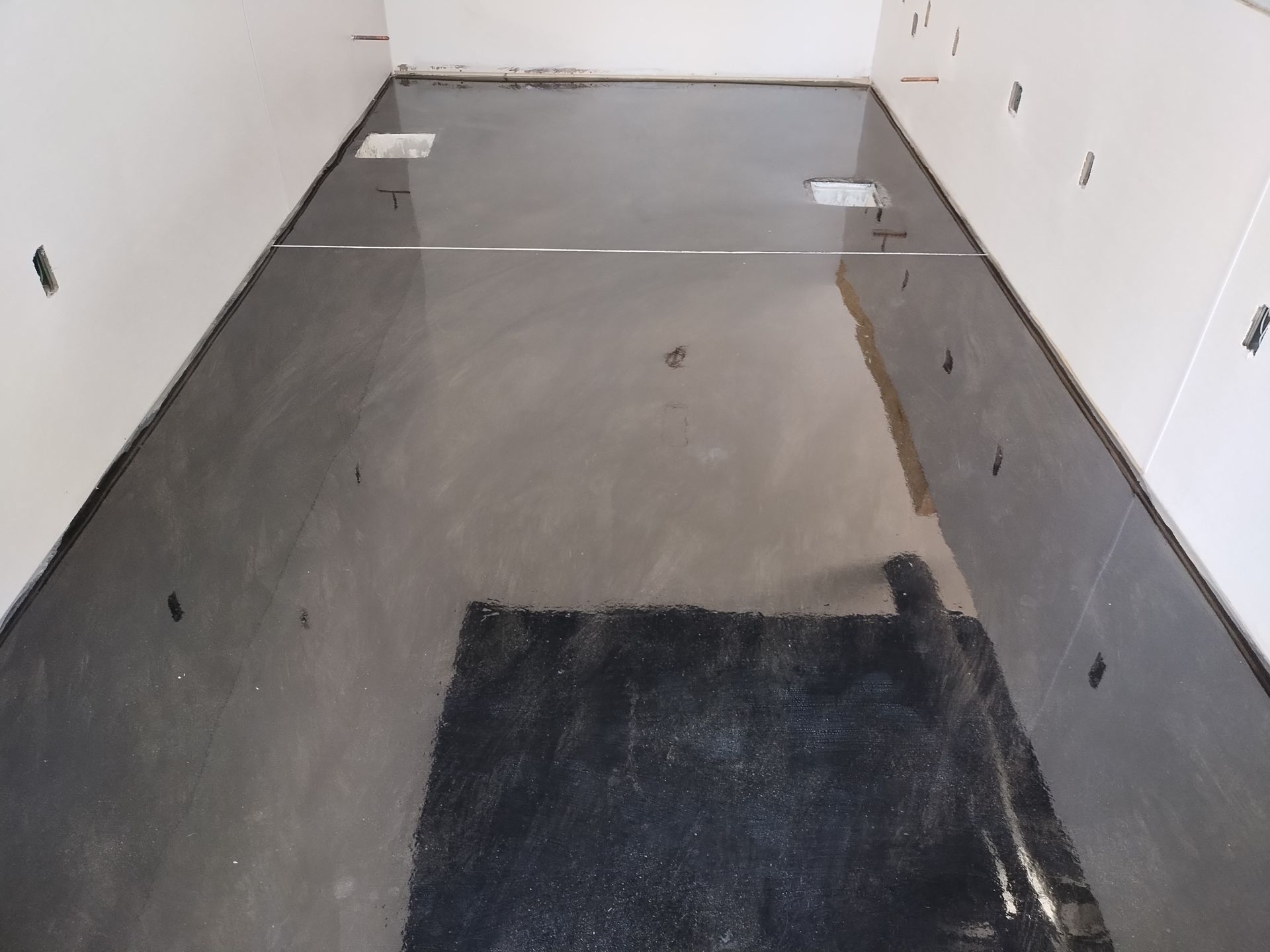 A smooth, light-gray concrete floor surface in a room with white walls and a dark rectangular floor mat in the foreground.