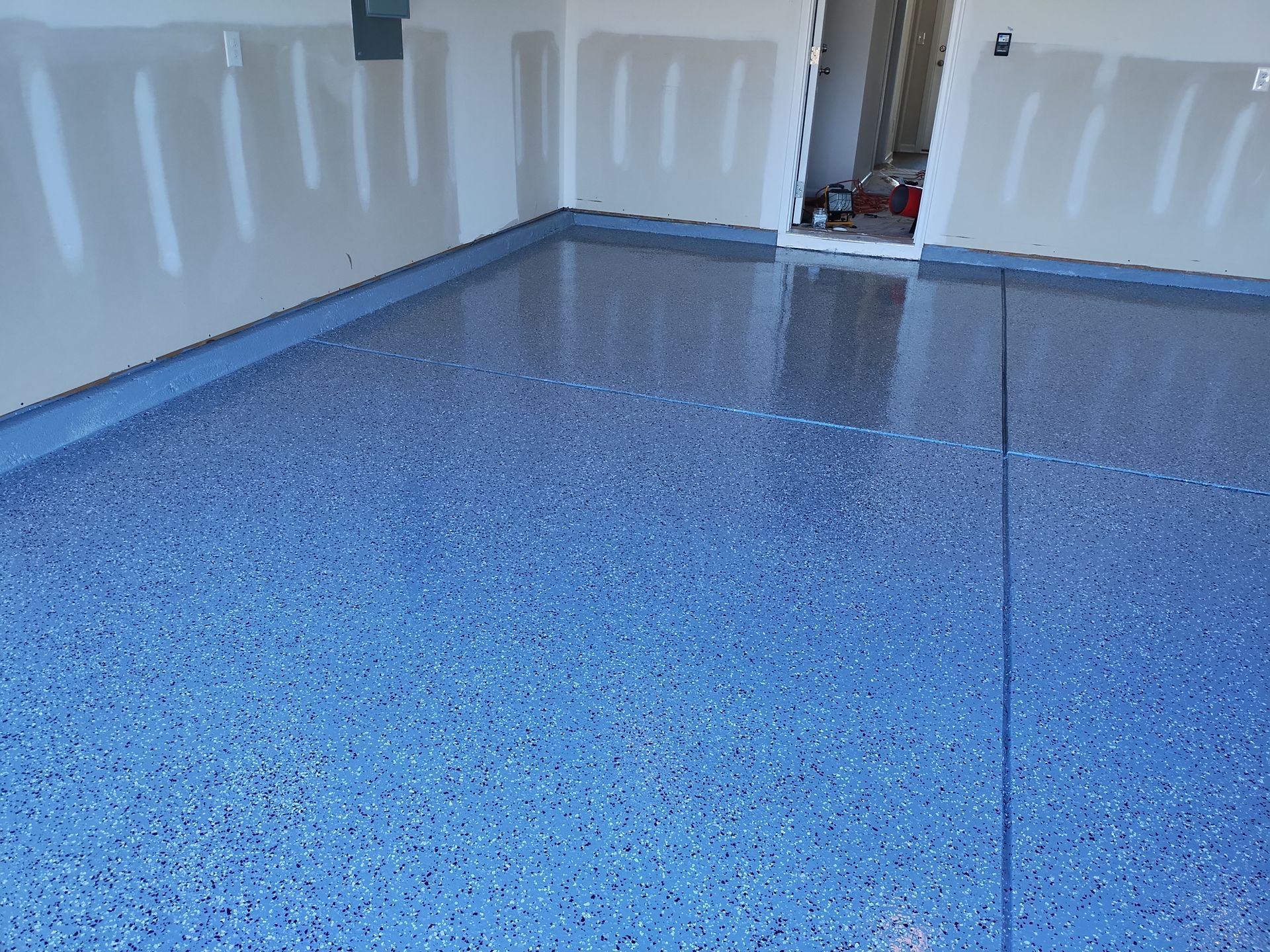 A glossy, blue-flecked epoxy floor in a garage with unfinished drywall walls and a doorway in the background.