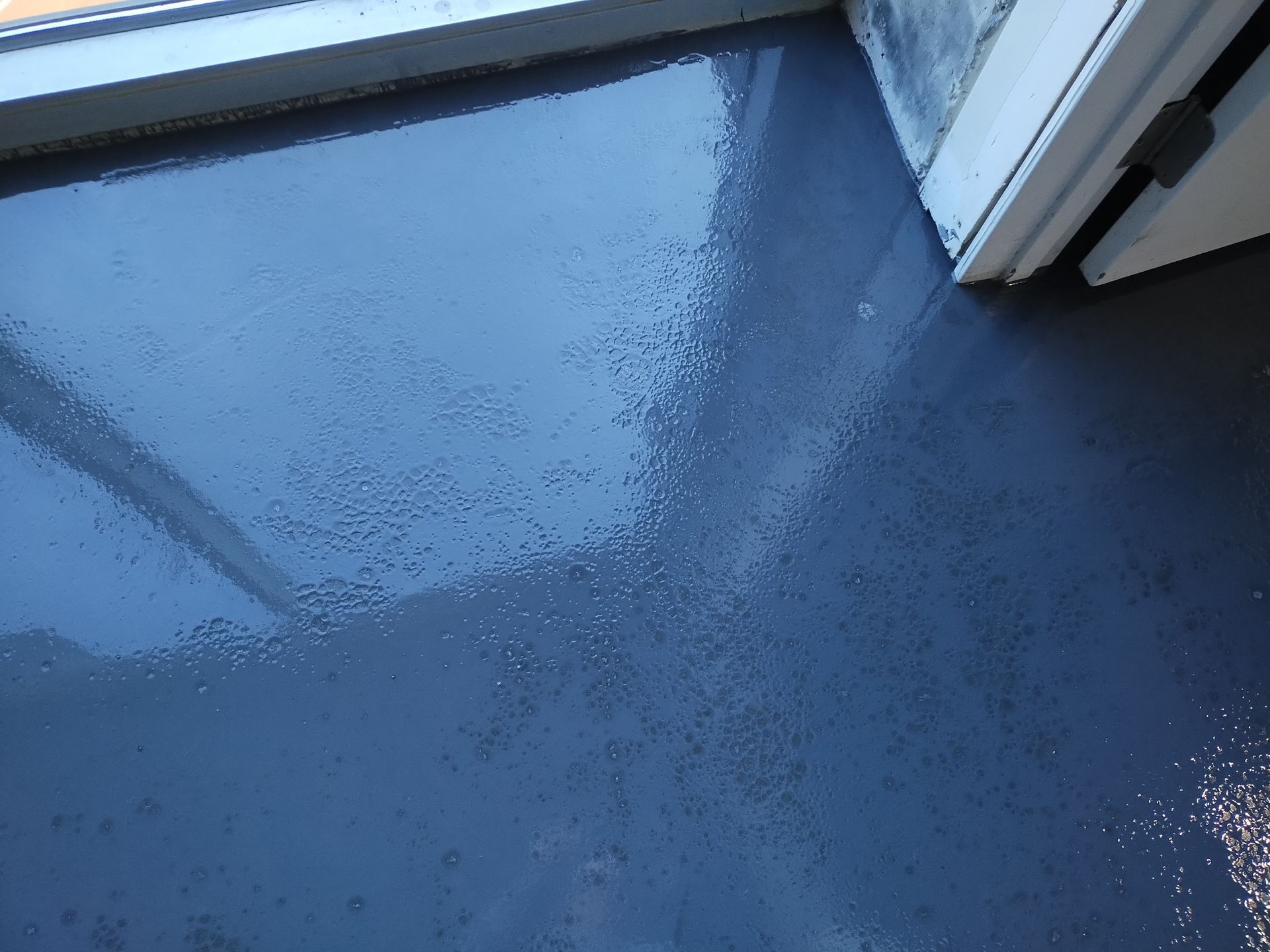 A close-up view of a recently applied, glossy blue epoxy floor finish with visible bubbles, near a doorway and wall.