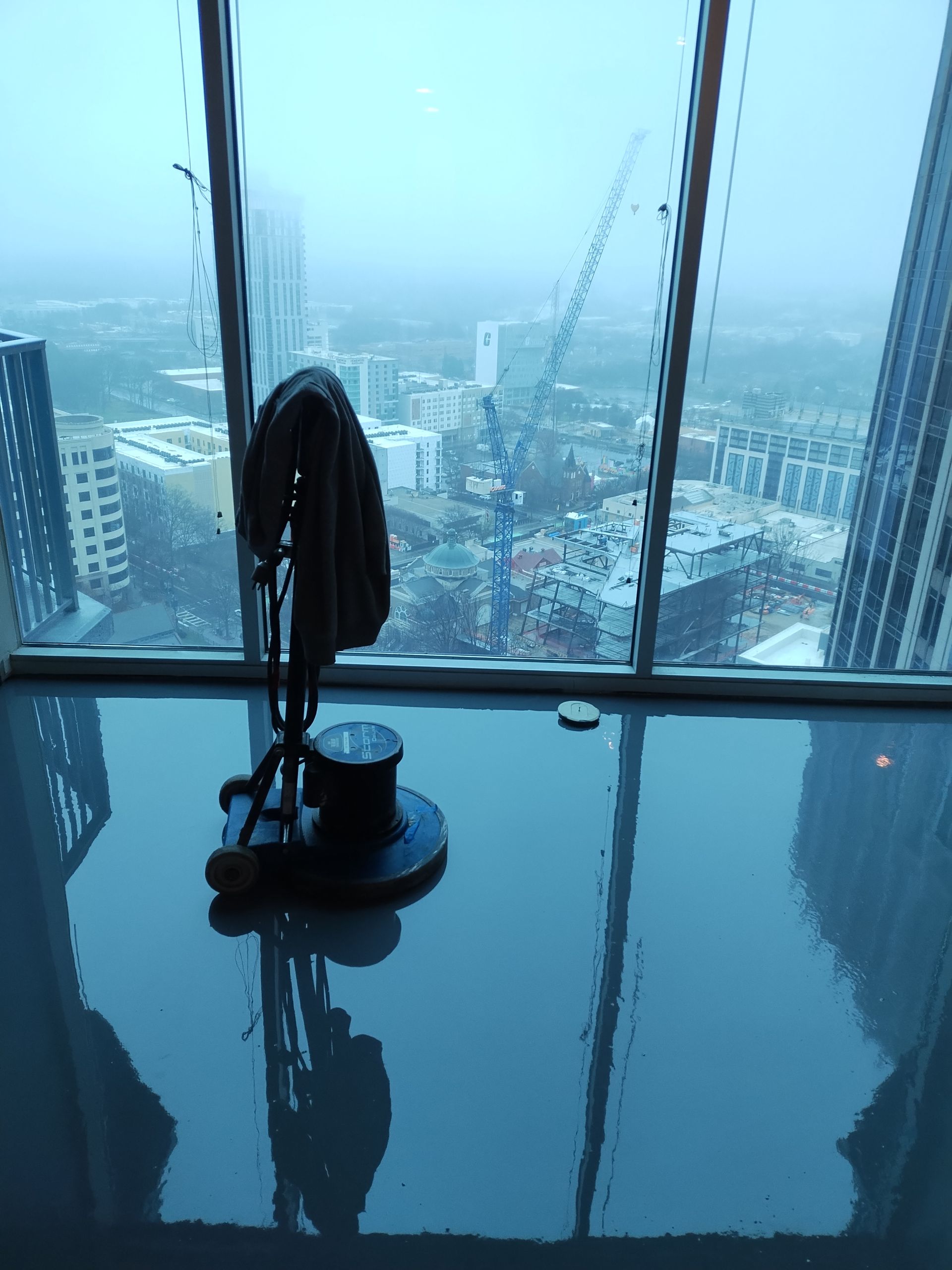 A floor buffer sits on a highly reflective, dark, polished floor in front of floor-to-ceiling windows overlooking a city.