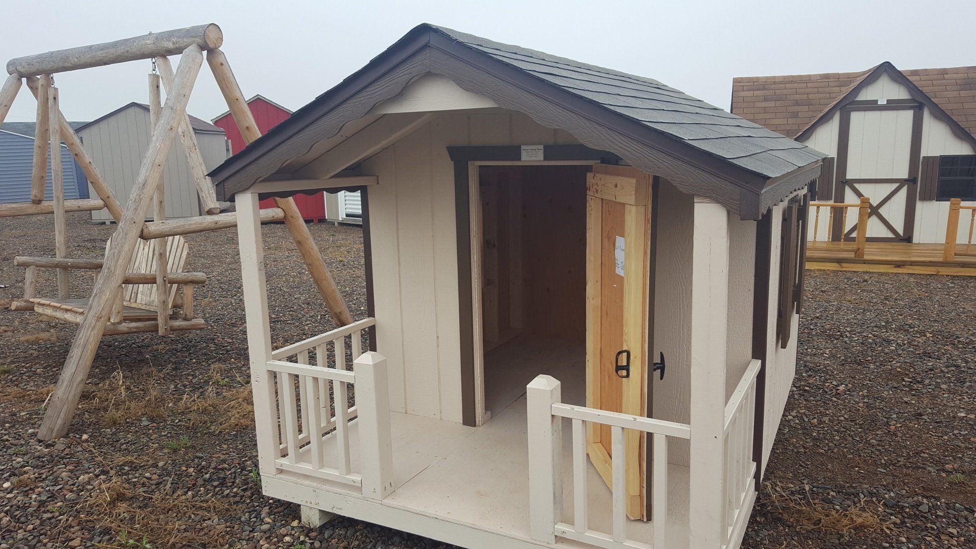 A small white playhouse with a porch and a swing set in the background.