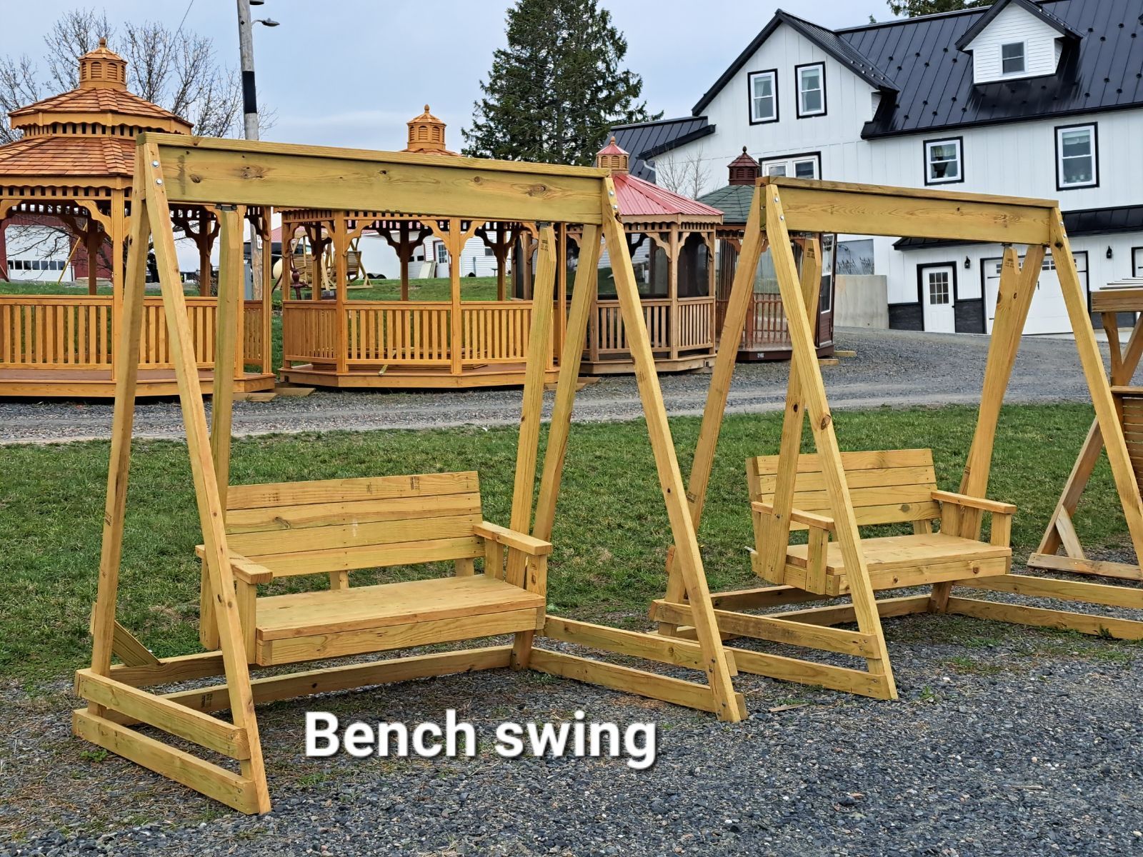 Two wooden bench swings are sitting next to each other in a gravel yard.