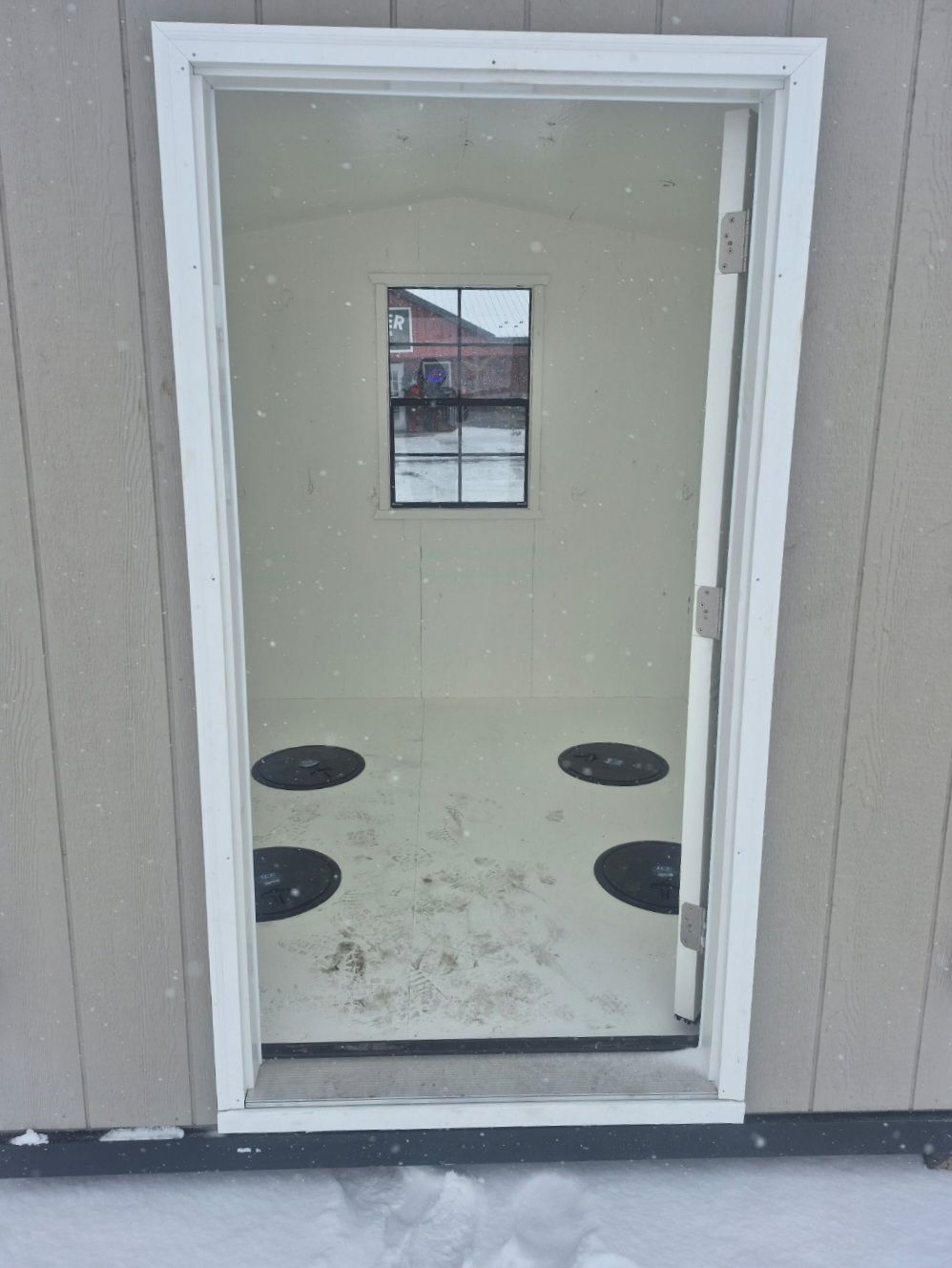 Interior of a beige ice fishing shack. Four dark circular holes in the white floor. Small window in the back wall.