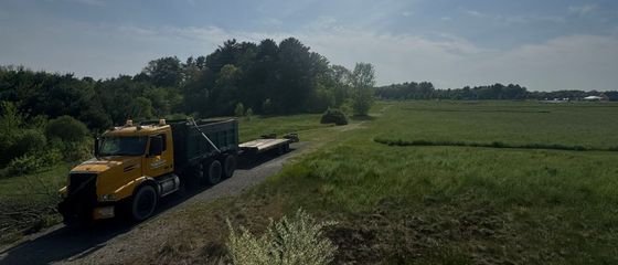 Yellow dump truck towing a flatbed trailer on a dirt road next to a field. Green trees and sky in background.