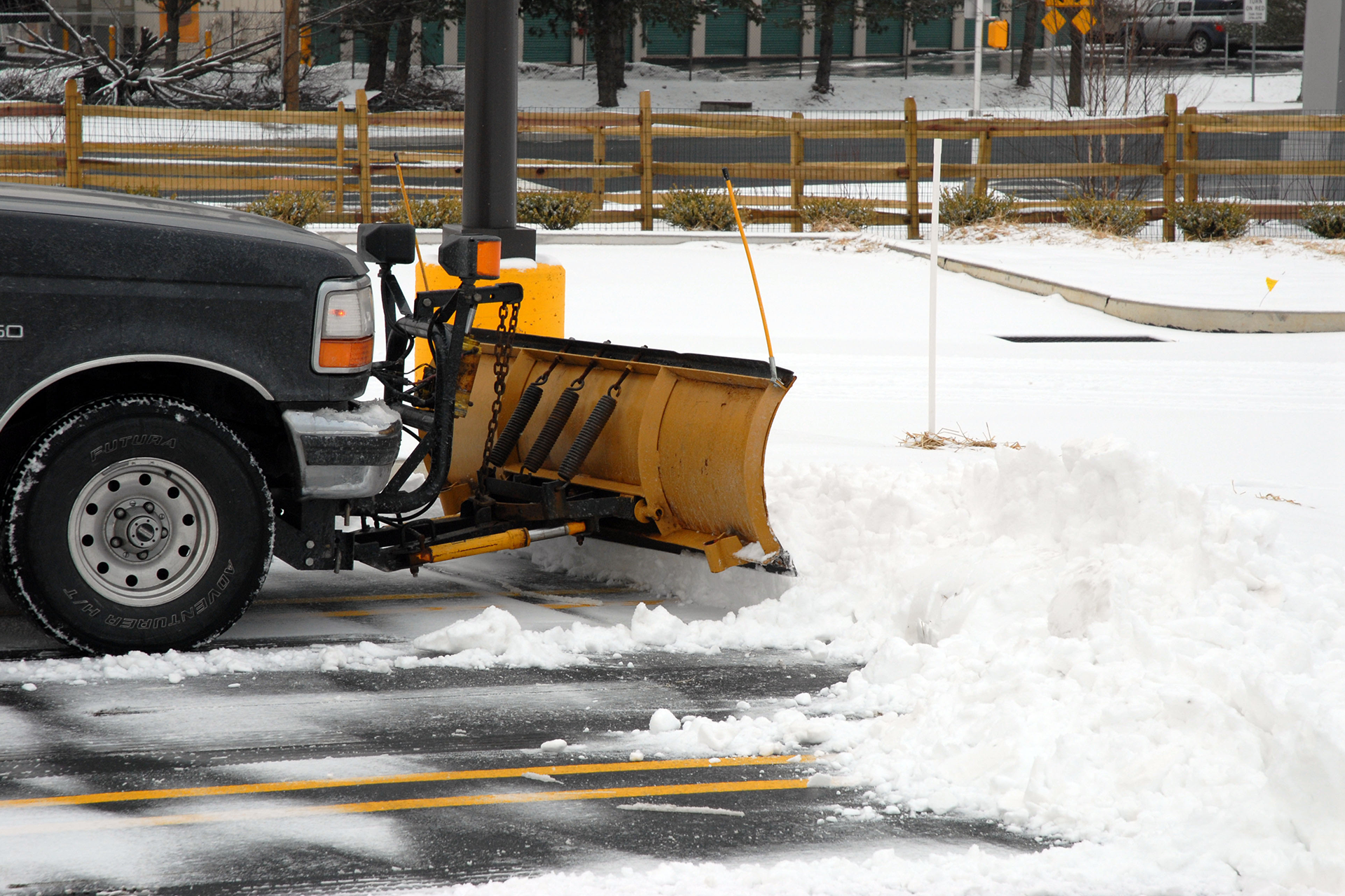 Snowplow clearing snow from a parking lot; vehicle is black with a yellow plow.