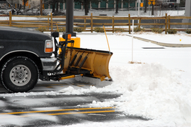 Snowplow clearing snow from a parking lot; vehicle is black with a yellow plow.