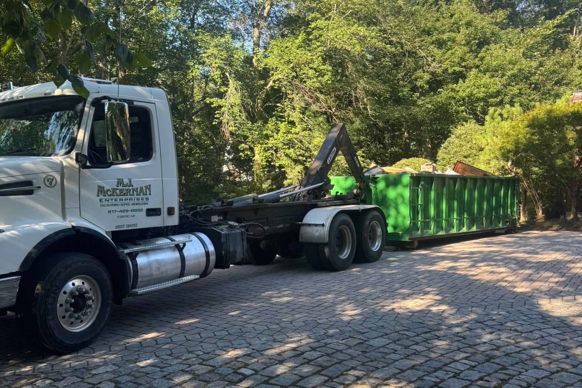 White truck with a green dumpster on a paved driveway; trees in the background.