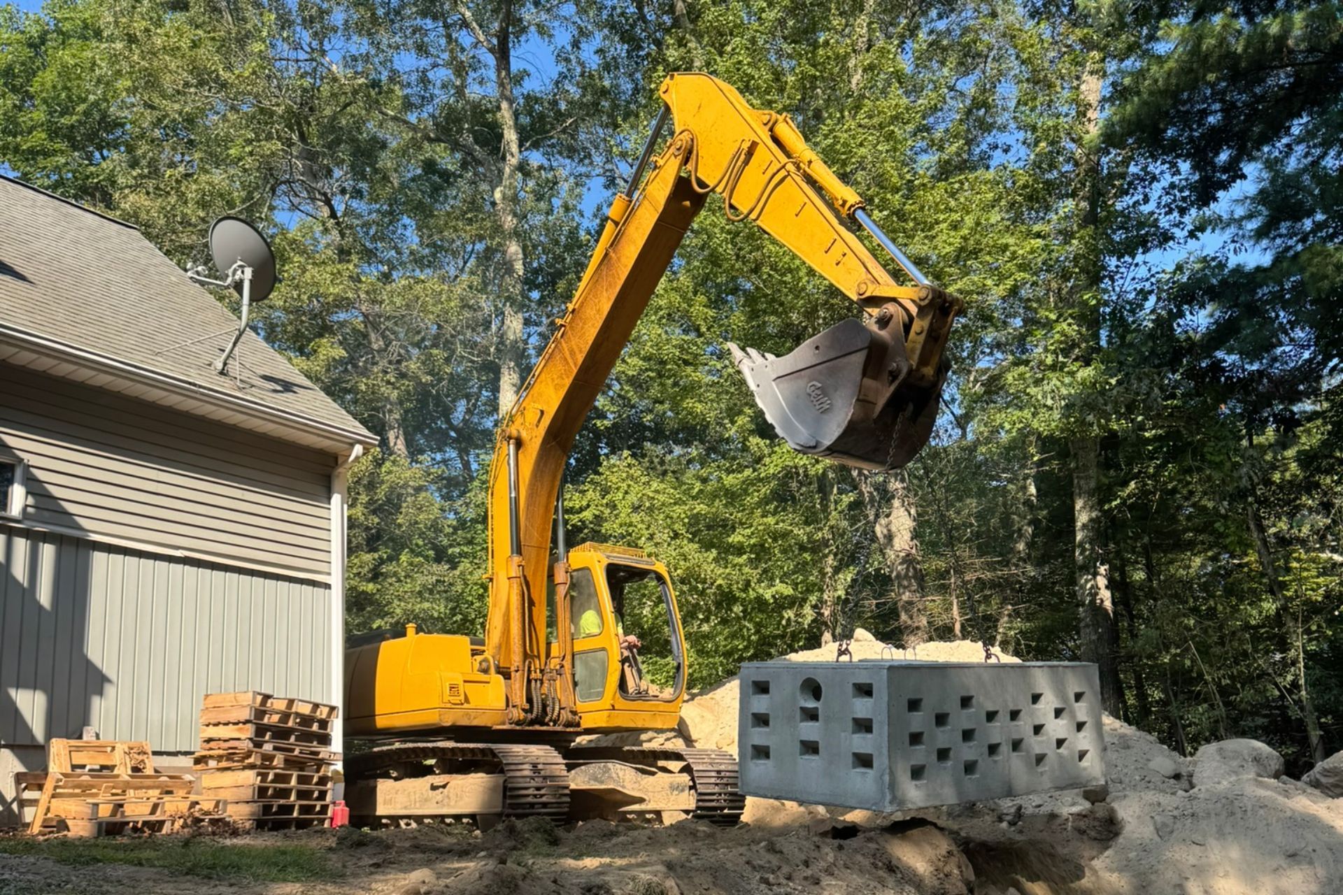 Yellow excavator lifting a large concrete block near a house with trees in the background.