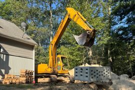 Yellow excavator lifting a large concrete block near a house with trees in the background.