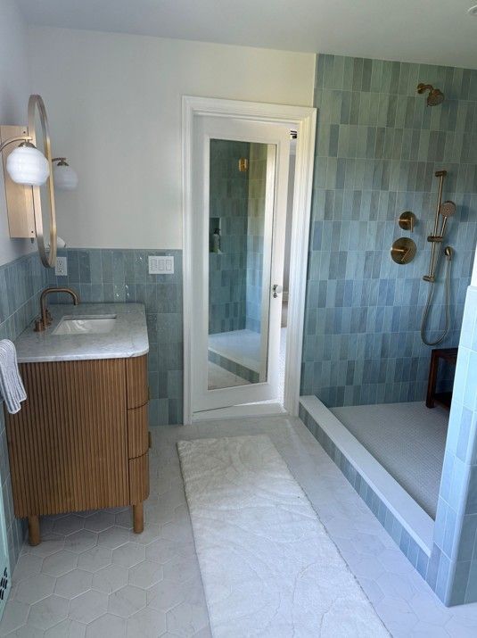 Bathroom with blue tiled shower, wood vanity, gold fixtures, and a white rug.