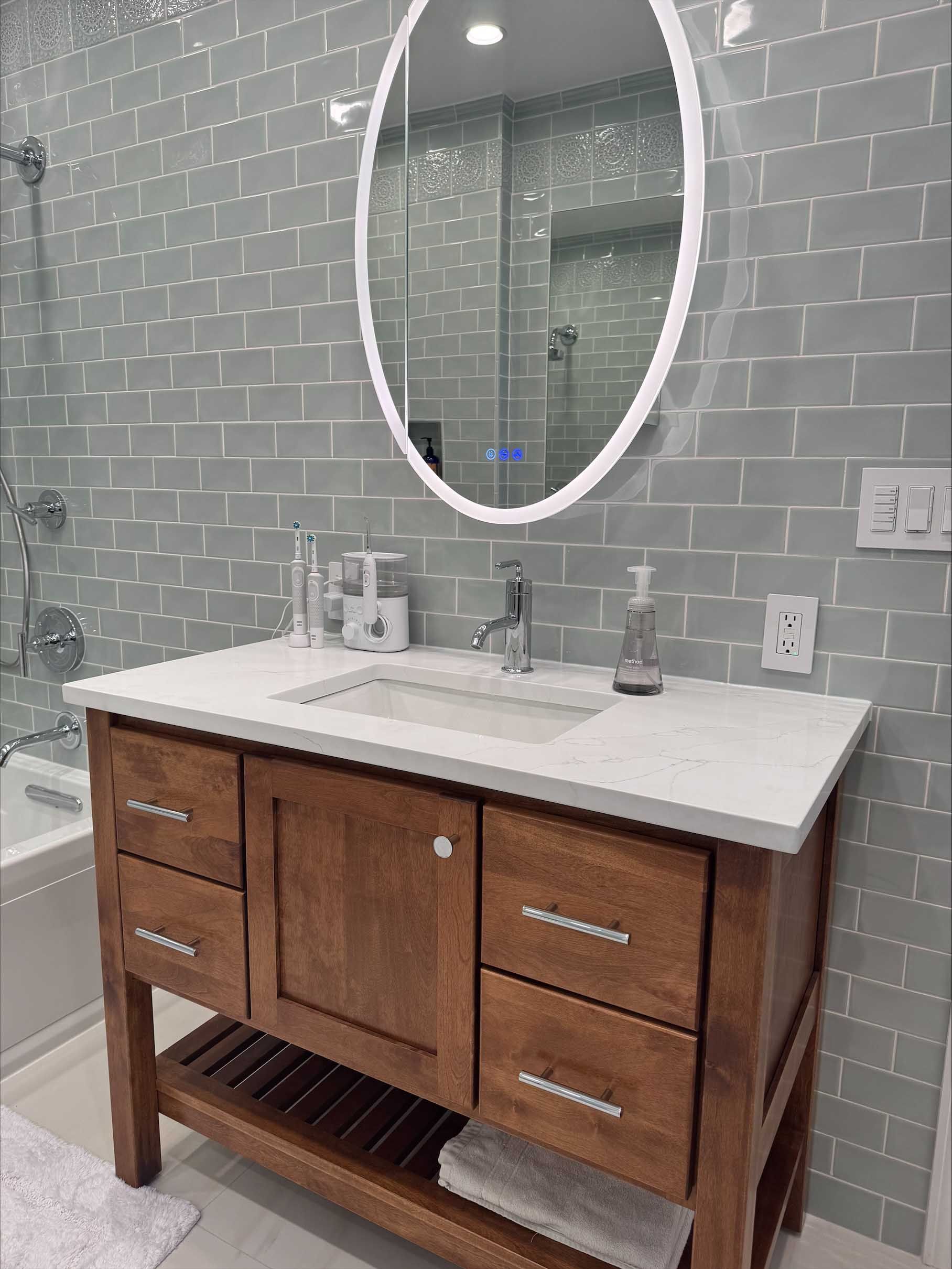 Bathroom vanity with a wood cabinet, white countertop, oval mirror, and gray tiled wall.