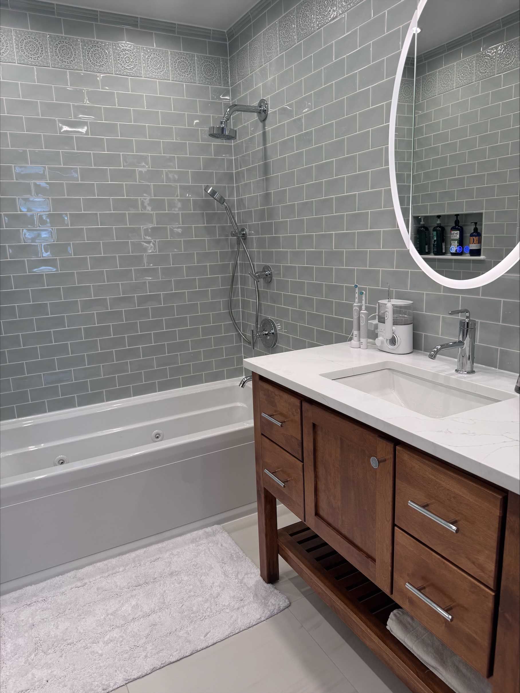 Bathroom with silver-tiled walls, a white tub, wooden vanity with a white countertop, and a circular mirror.