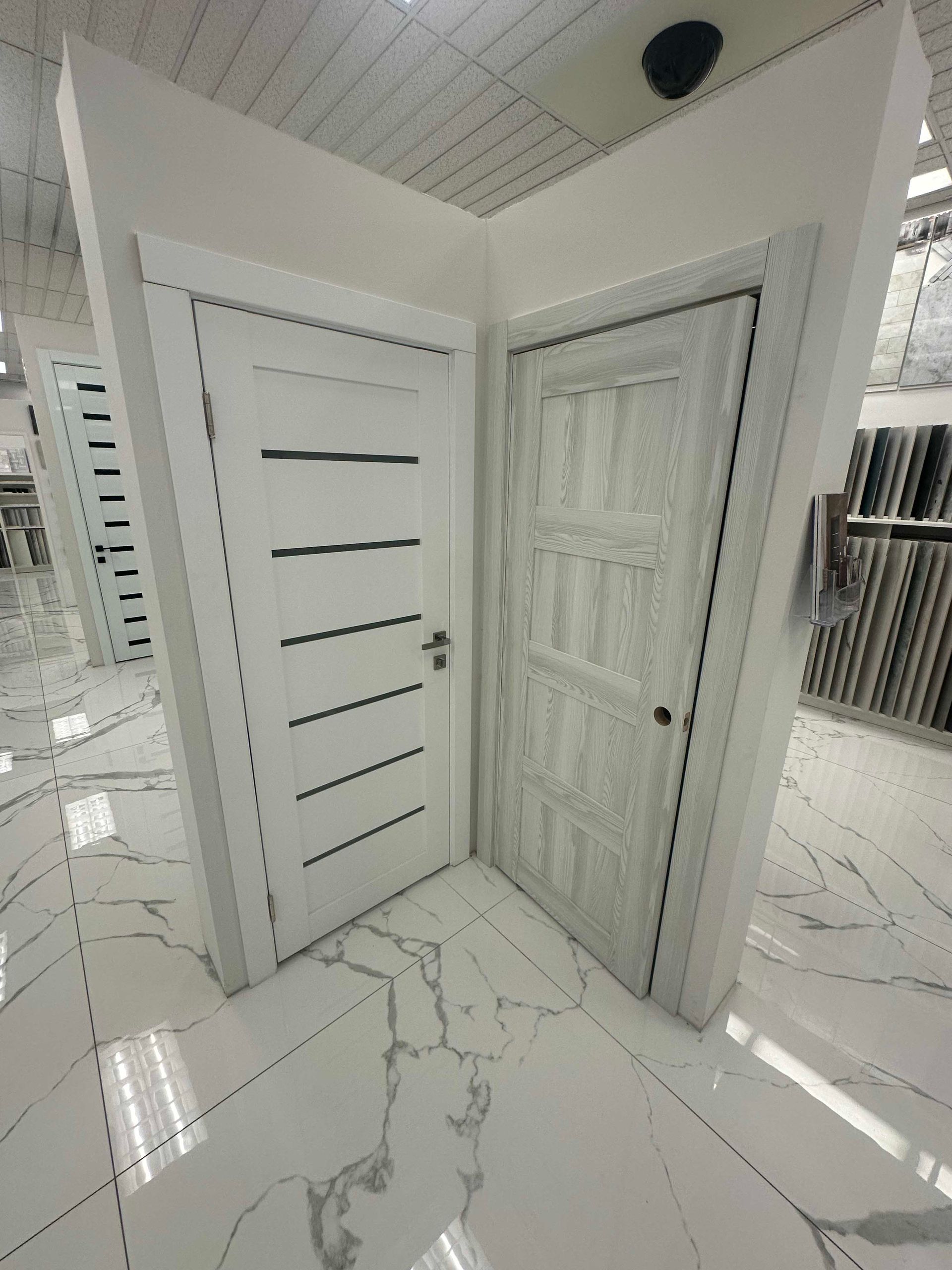 Two white interior doors displayed in a showroom. The door on the left has horizontal black lines. The floor is white marble.