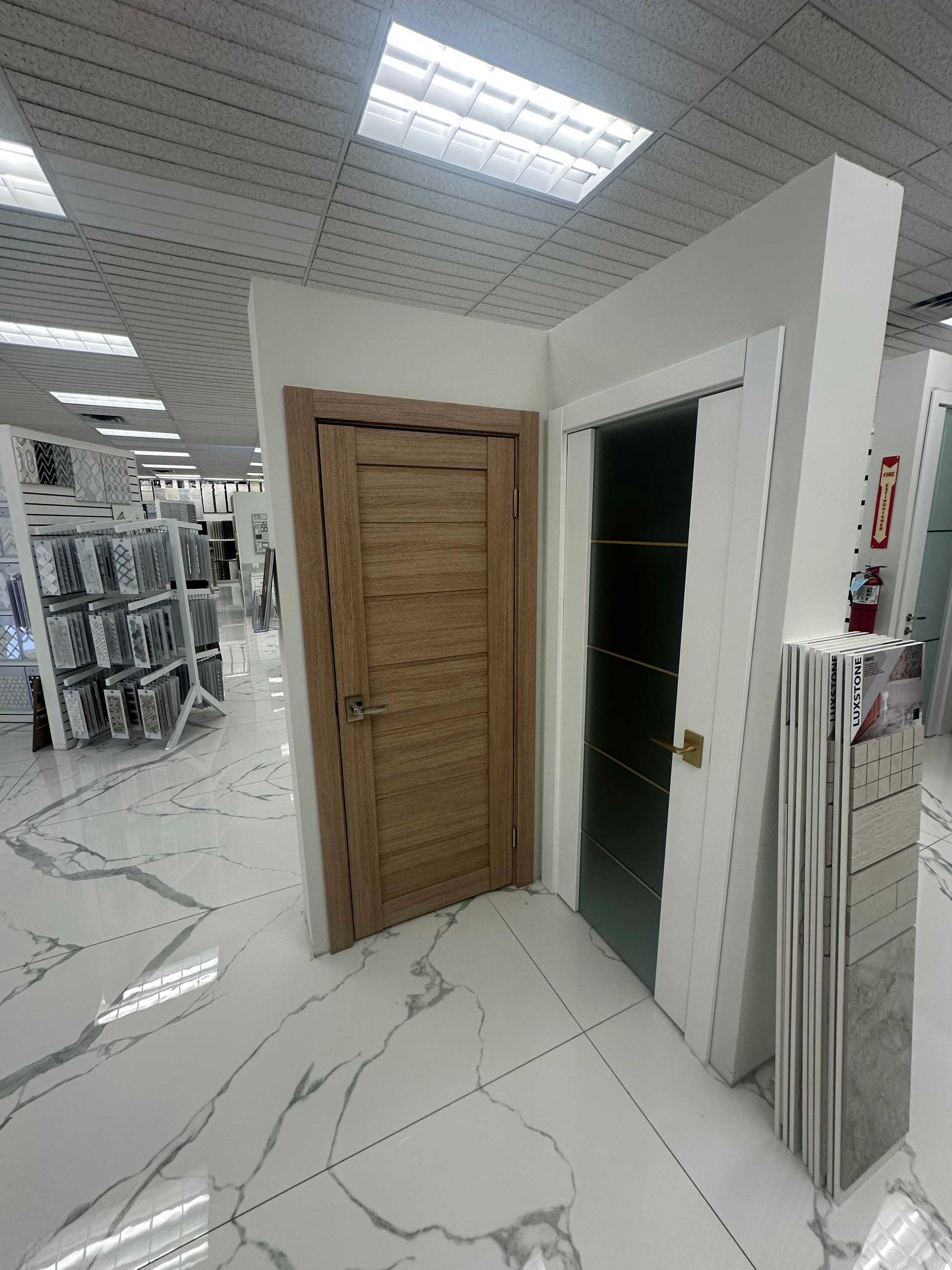 Two interior doors displayed in a showroom. One is wooden, the other glass. White walls and marble tile floors.