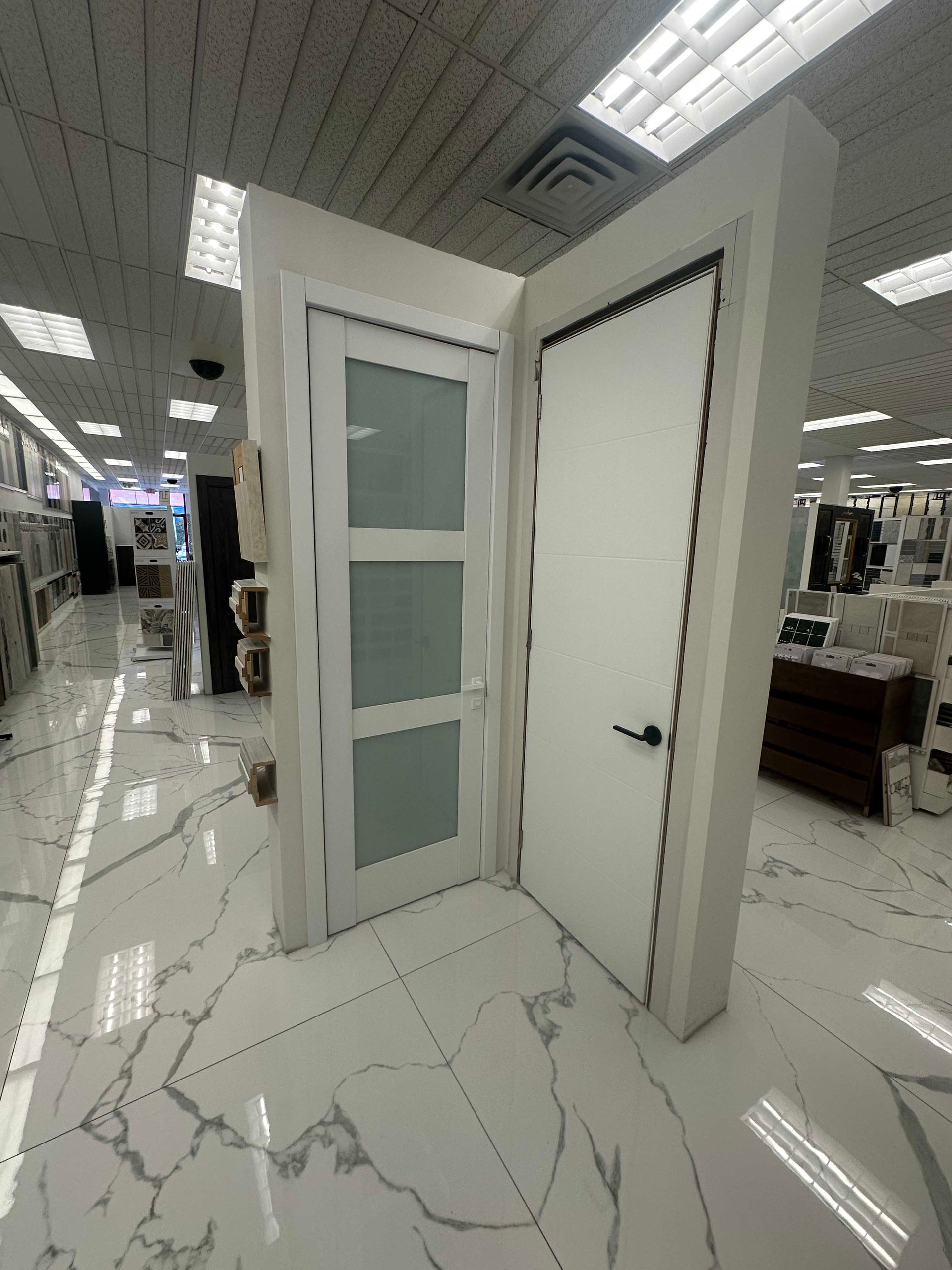 Two white interior doors displayed in a showroom with white marble tile flooring and overhead lighting.