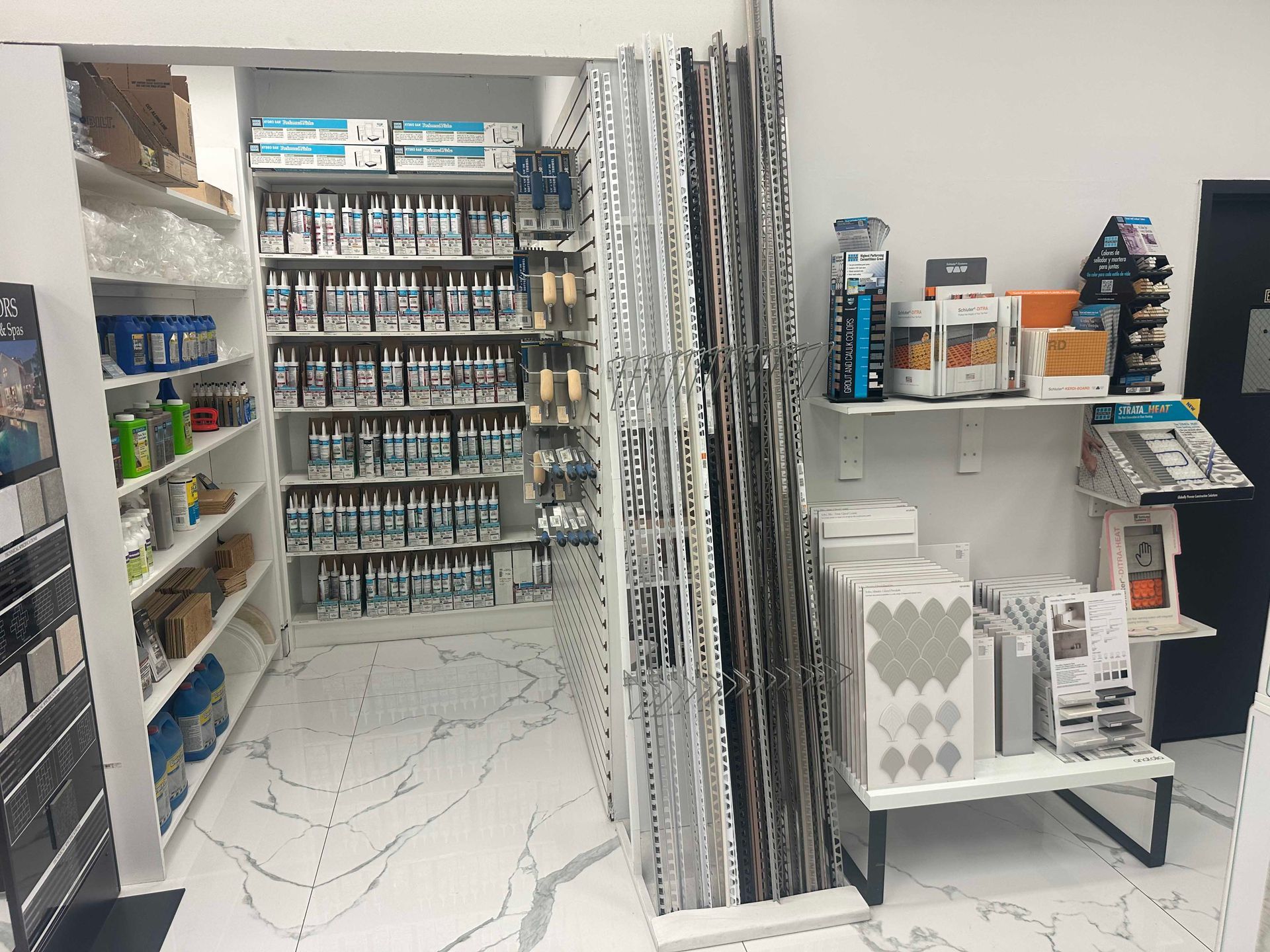 Interior of a retail store displays shelves stocked with various construction supplies like caulk, tile samples, and trim.