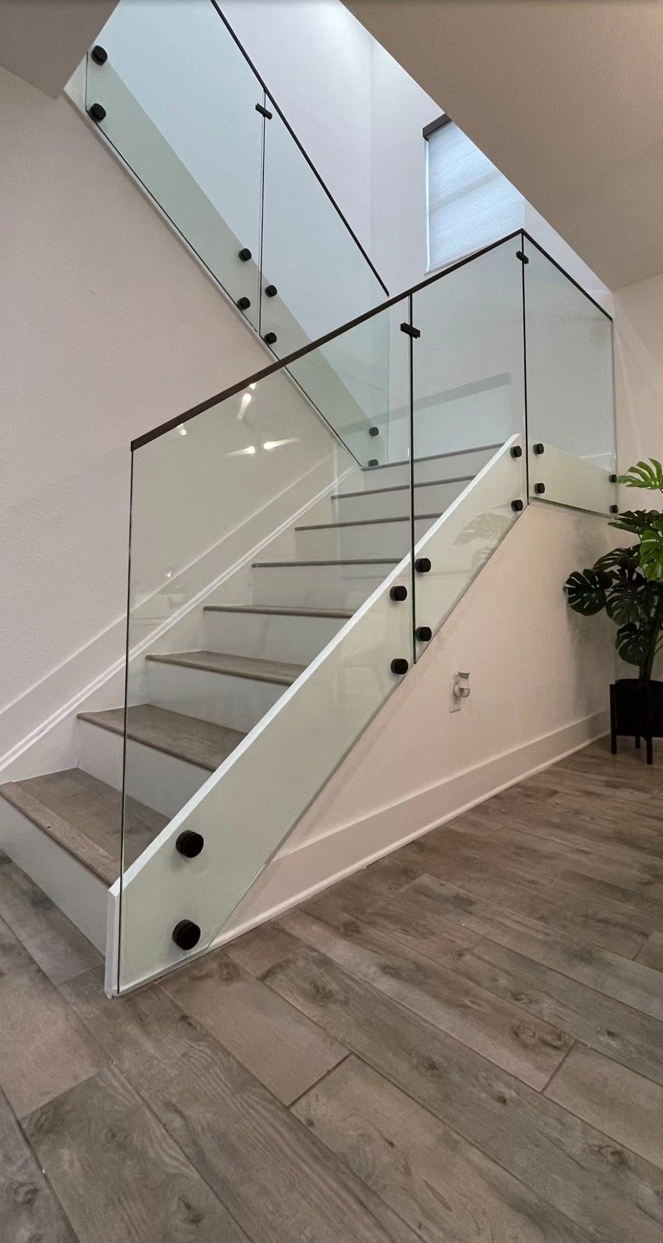 Staircase with glass railing and black hardware, white walls, and wood-look flooring.