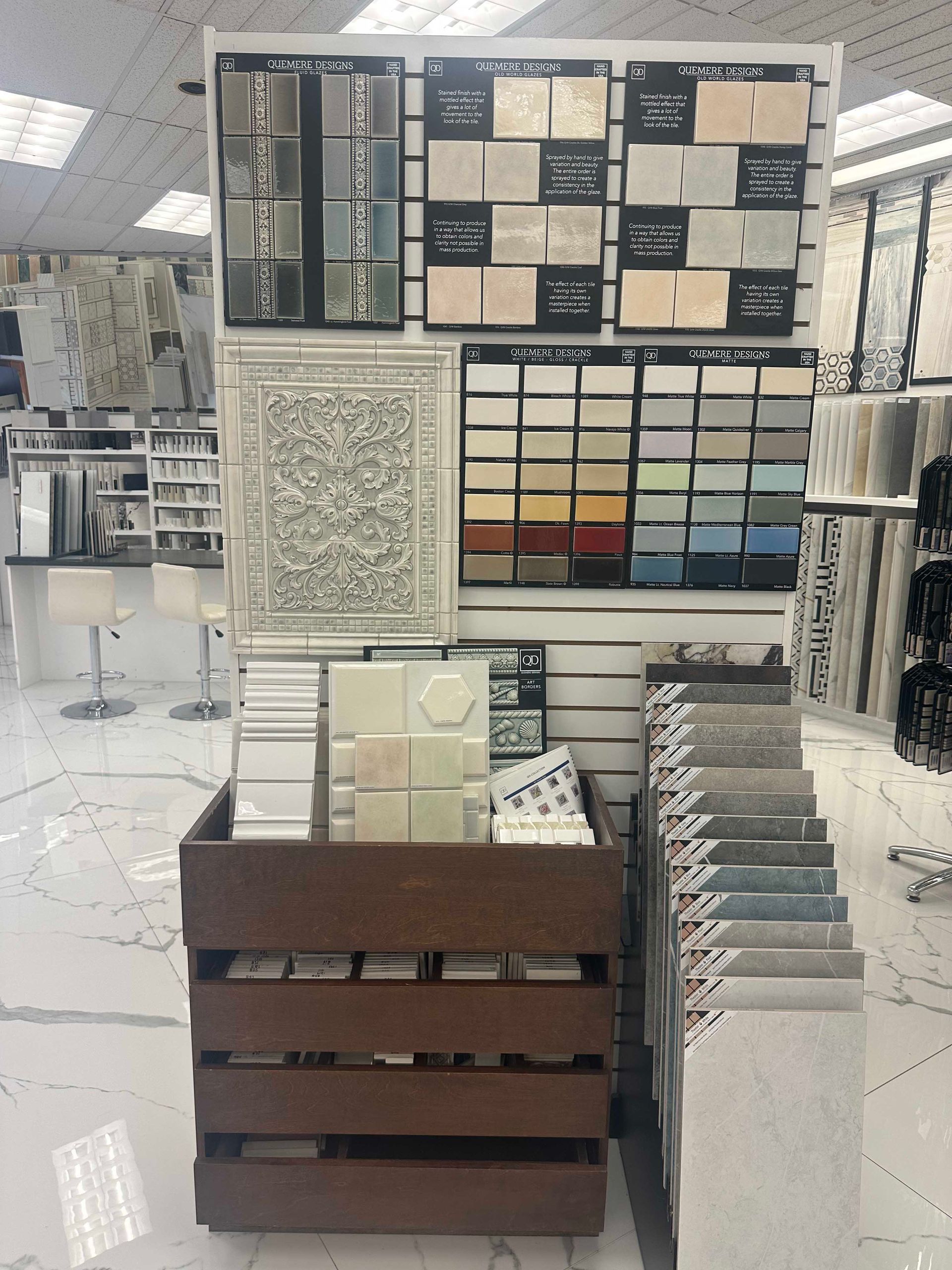 Tile samples on display in a retail setting, featuring various colors and patterns. A wooden crate holds additional samples.