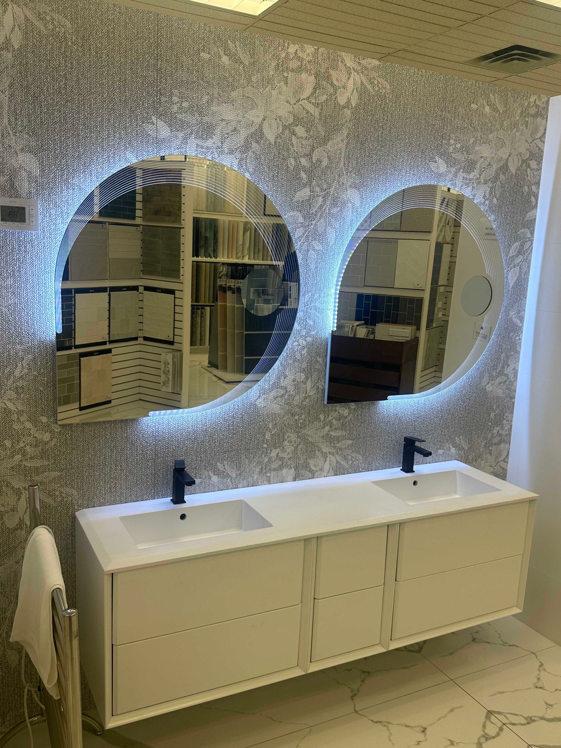 White floating vanity with dual sinks, two oval mirrors with backlighting, against textured wall.