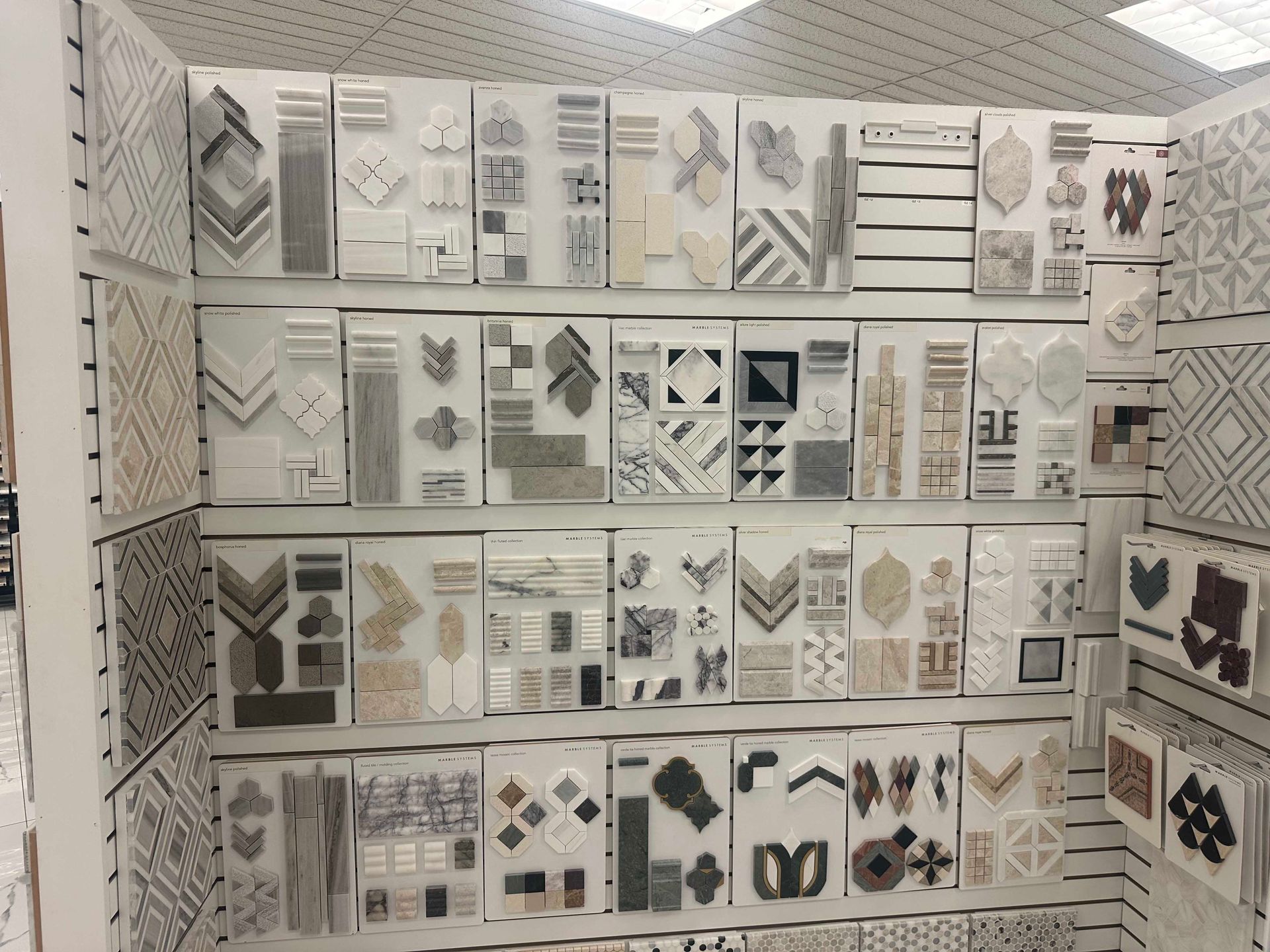 Tile samples displayed on white shelving in a store. Various shapes, patterns, and colors are visible.