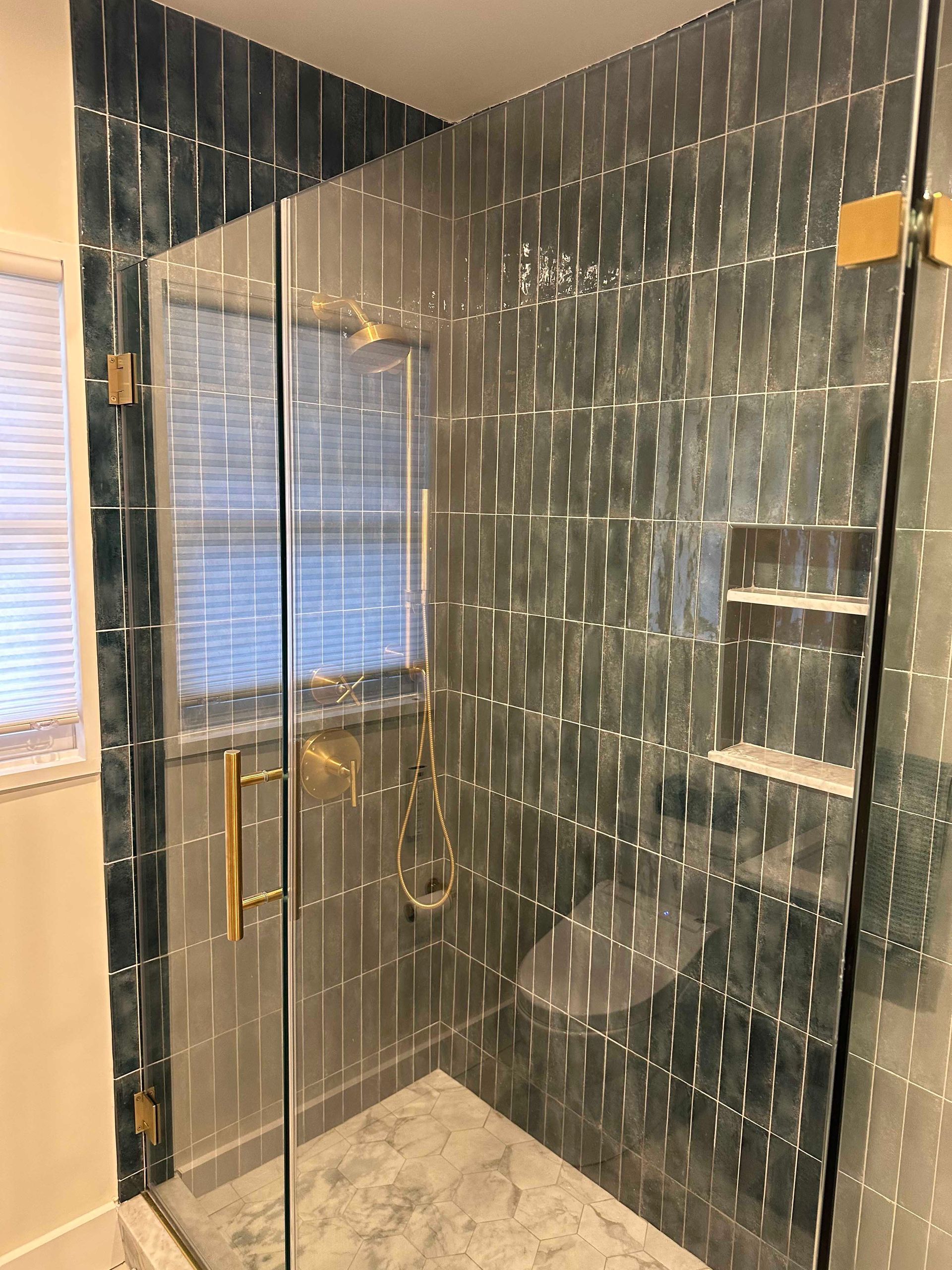 Shower with teal, textured tiles, glass door with gold hardware, and a window.