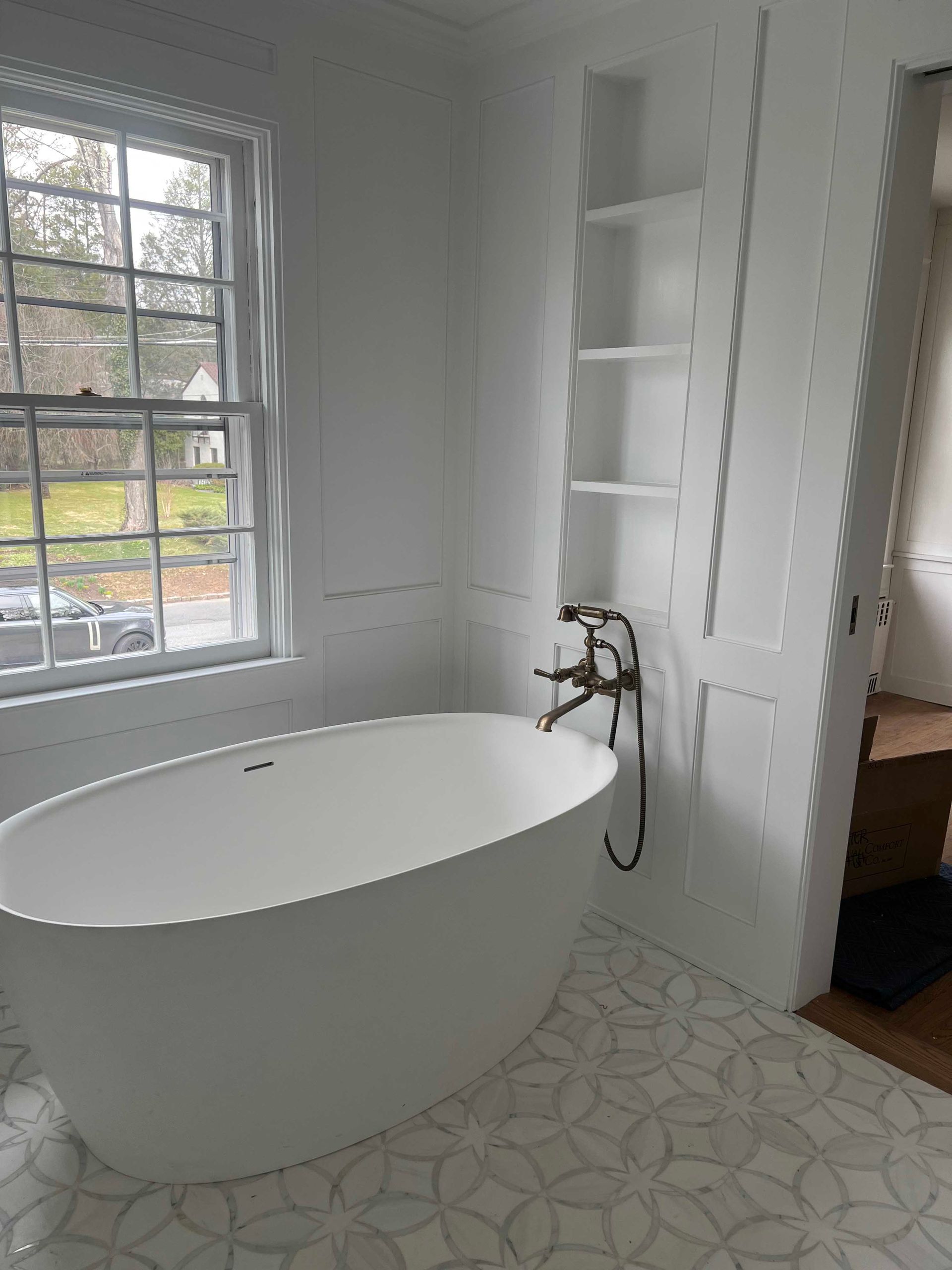 White bathroom with a freestanding tub, window, built-in shelves, and decorative floor tiles.