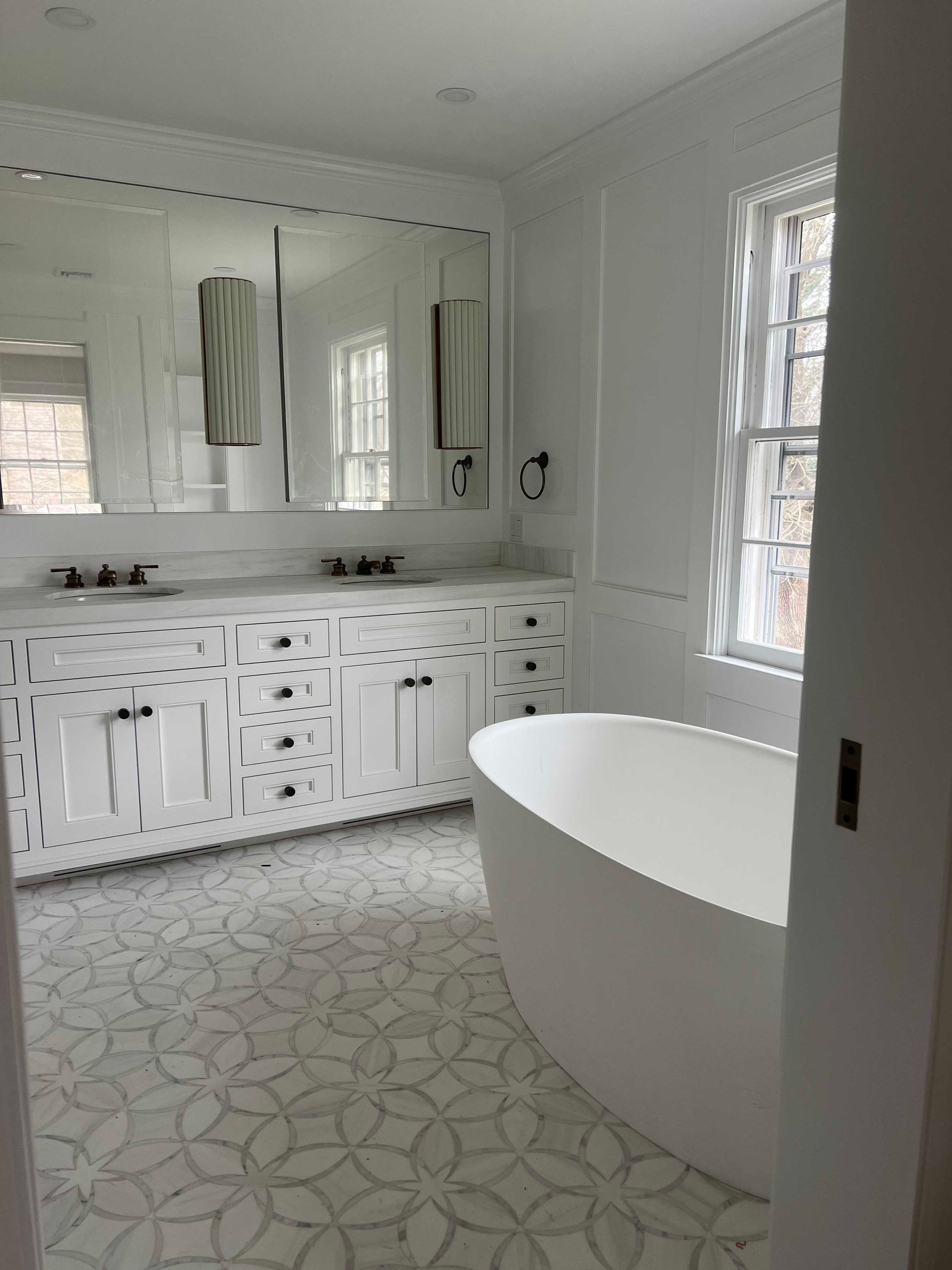 White bathroom with double vanity, free-standing tub, and decorative tile flooring.