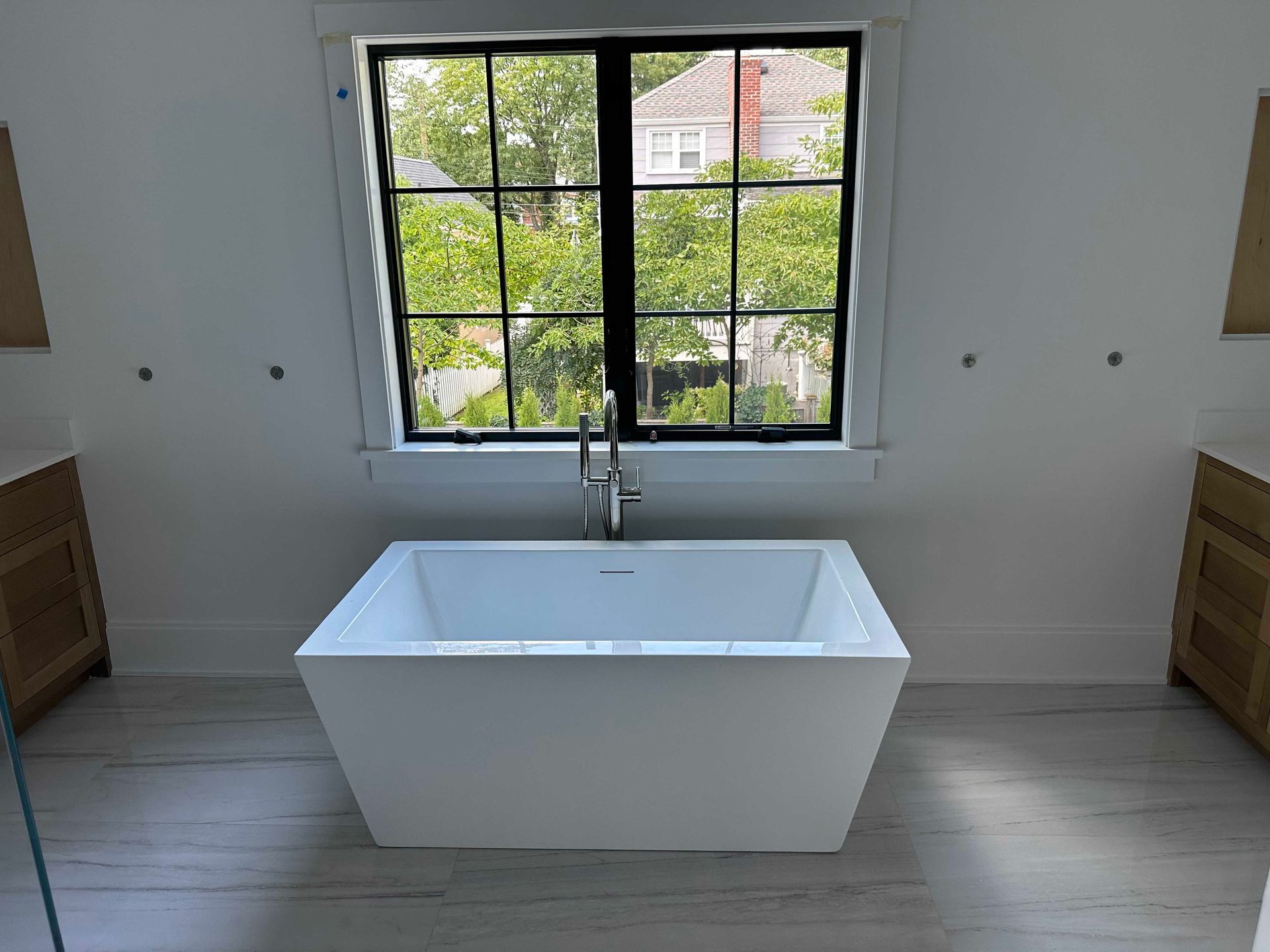 Modern white bathtub centered under a black-framed window. Light wood floor and two light-colored cabinets.