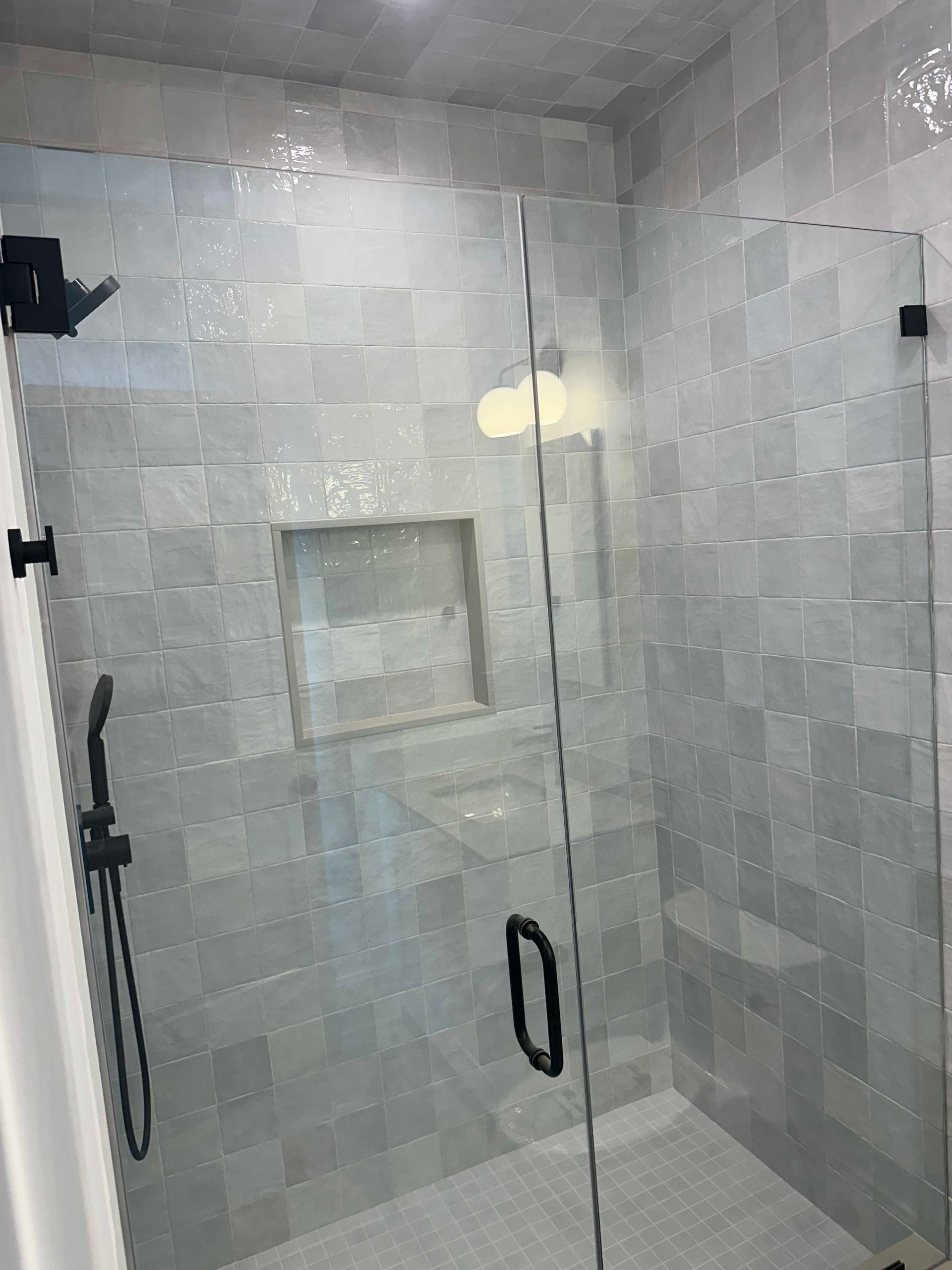 Shower with gray square tiles, glass door, black hardware, and built-in shelf.