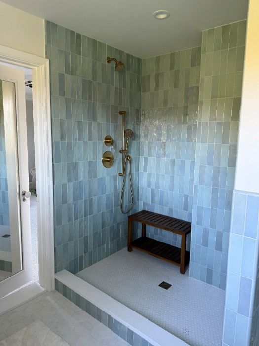 Walk-in shower with light blue tile walls, gold fixtures, a wooden bench, and white tile floor.