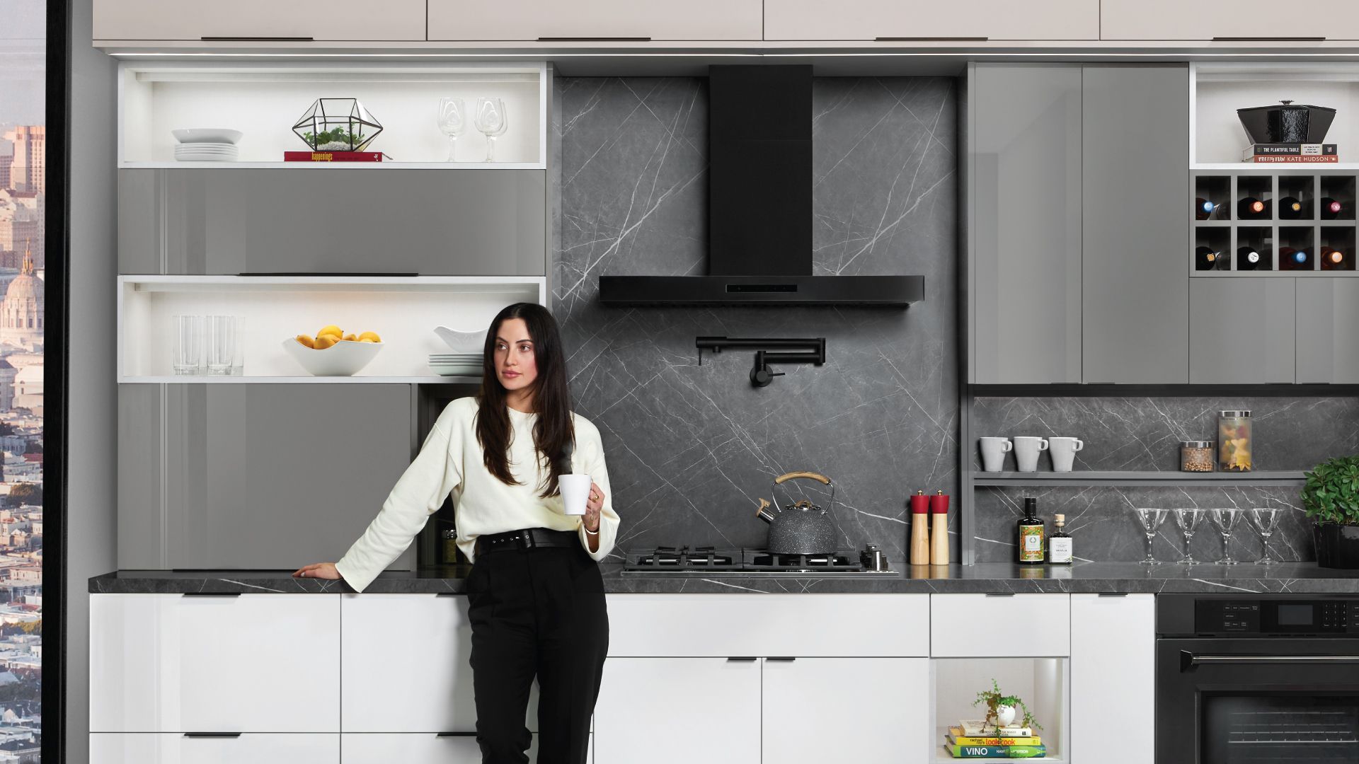 Woman in kitchen with white and gray cabinets, black range hood, and marble backsplash.