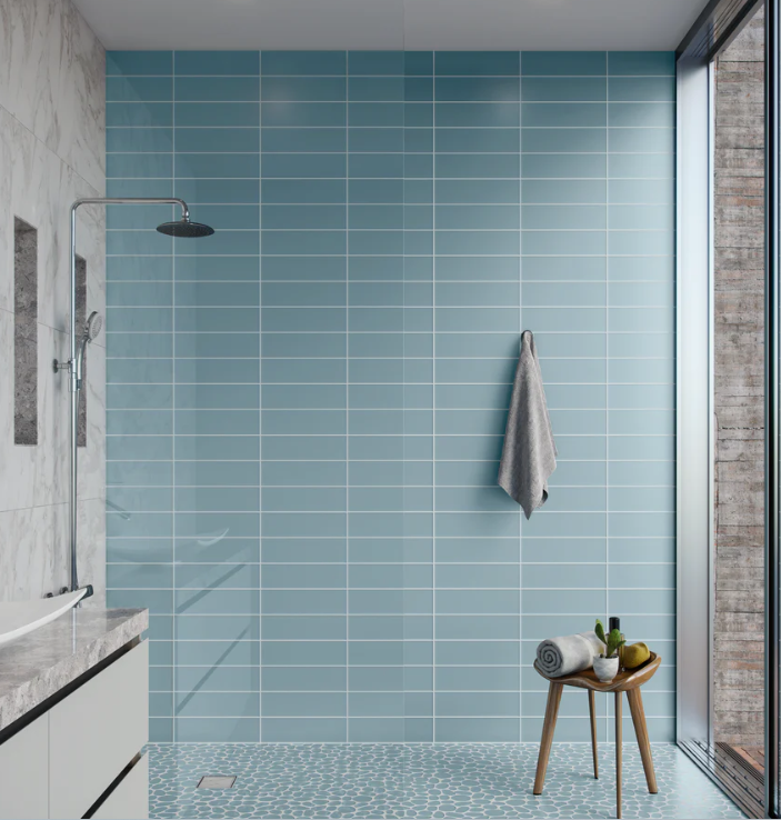Blue tiled shower with a towel hanging and a small wooden stool with items near a large window.