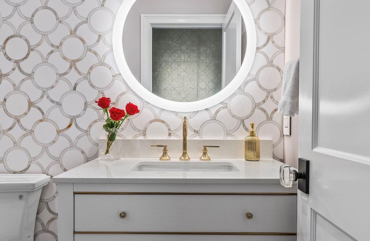 Bathroom vanity with gold fixtures, round mirror, patterned tile backsplash, and red roses.