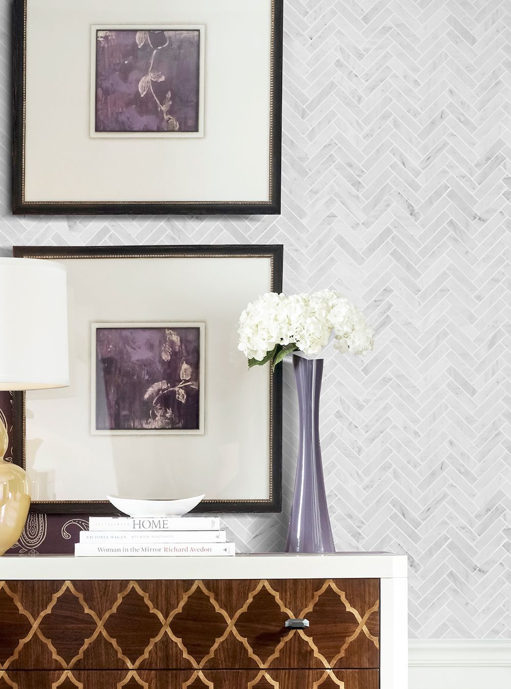 Two framed prints above a decorative cabinet with a vase of white flowers, against a herringbone-patterned wall.