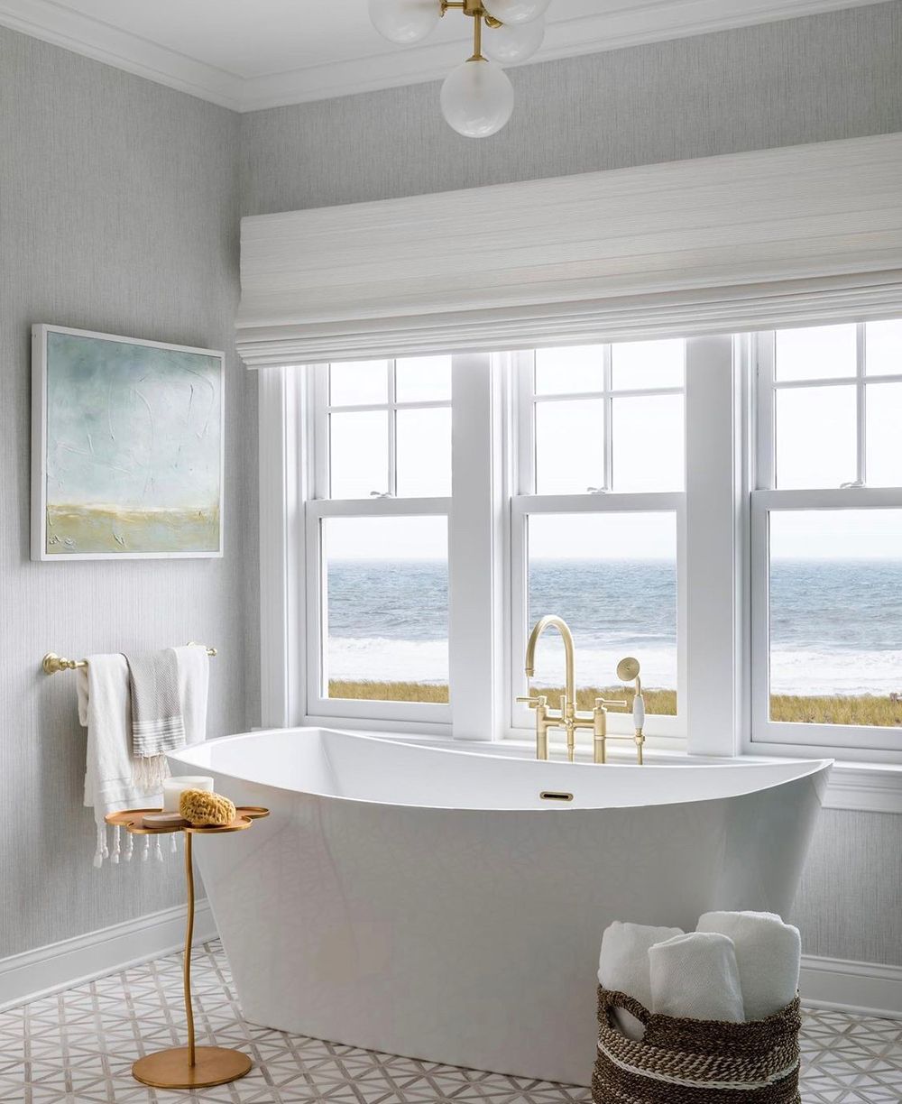 Bathroom with a white tub, large windows with ocean view, and gold fixtures.